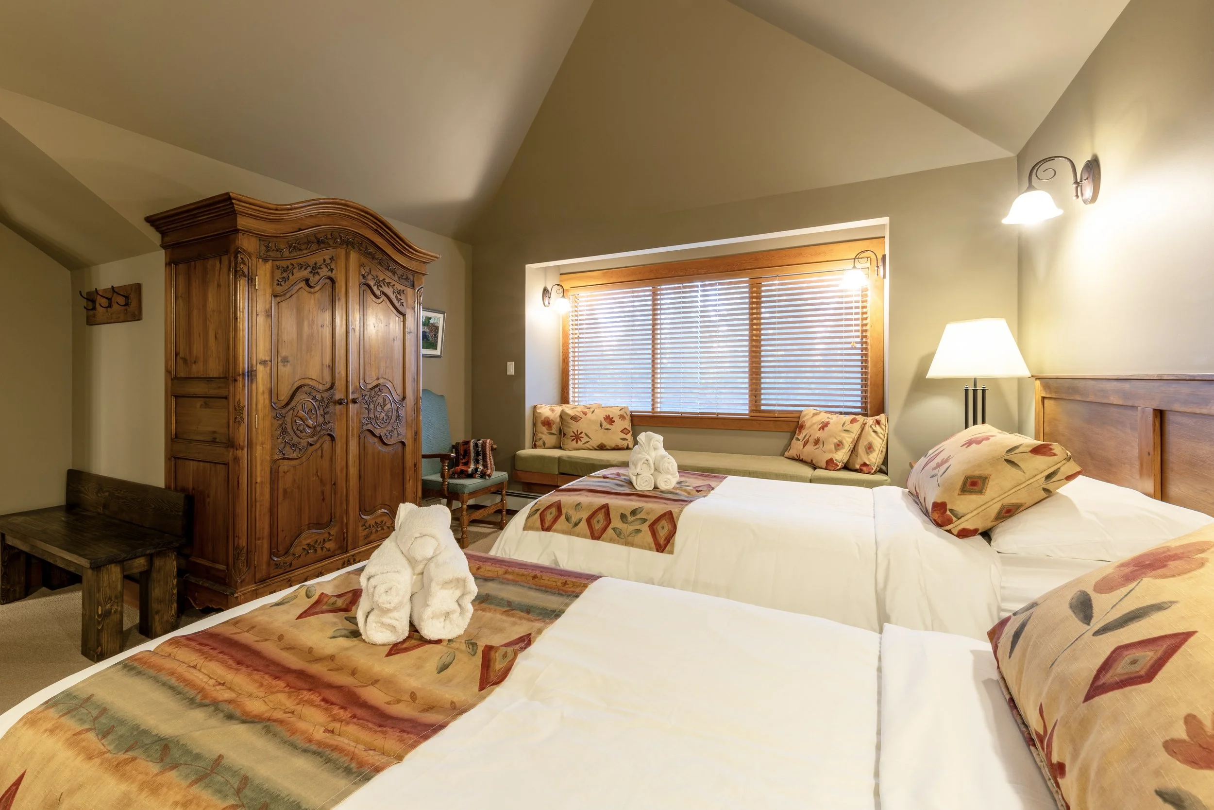 A cozy bedroom with two beds, a wooden wardrobe, a window with blinds, a seating area with cushions, a table with towel art, and warm lighting.