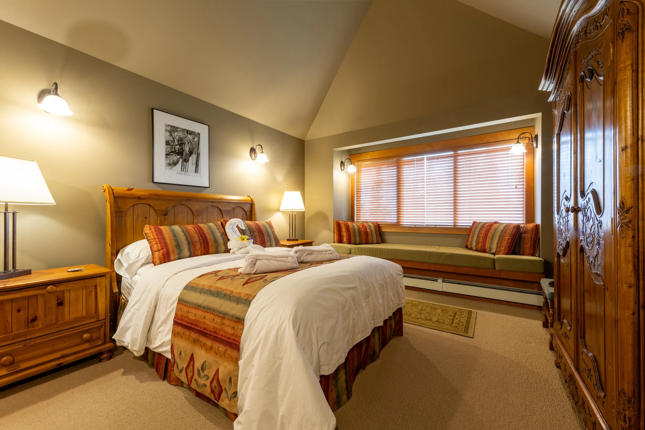 A cozy hotel room with a large bed, wooden furniture, and warm lighting. The bed has a swan towel decoration and colorful pillows. There's a large window with wooden blinds and a built-in bench beneath it. The room features beige walls, a framed picture of a moose, and matching wall sconces.