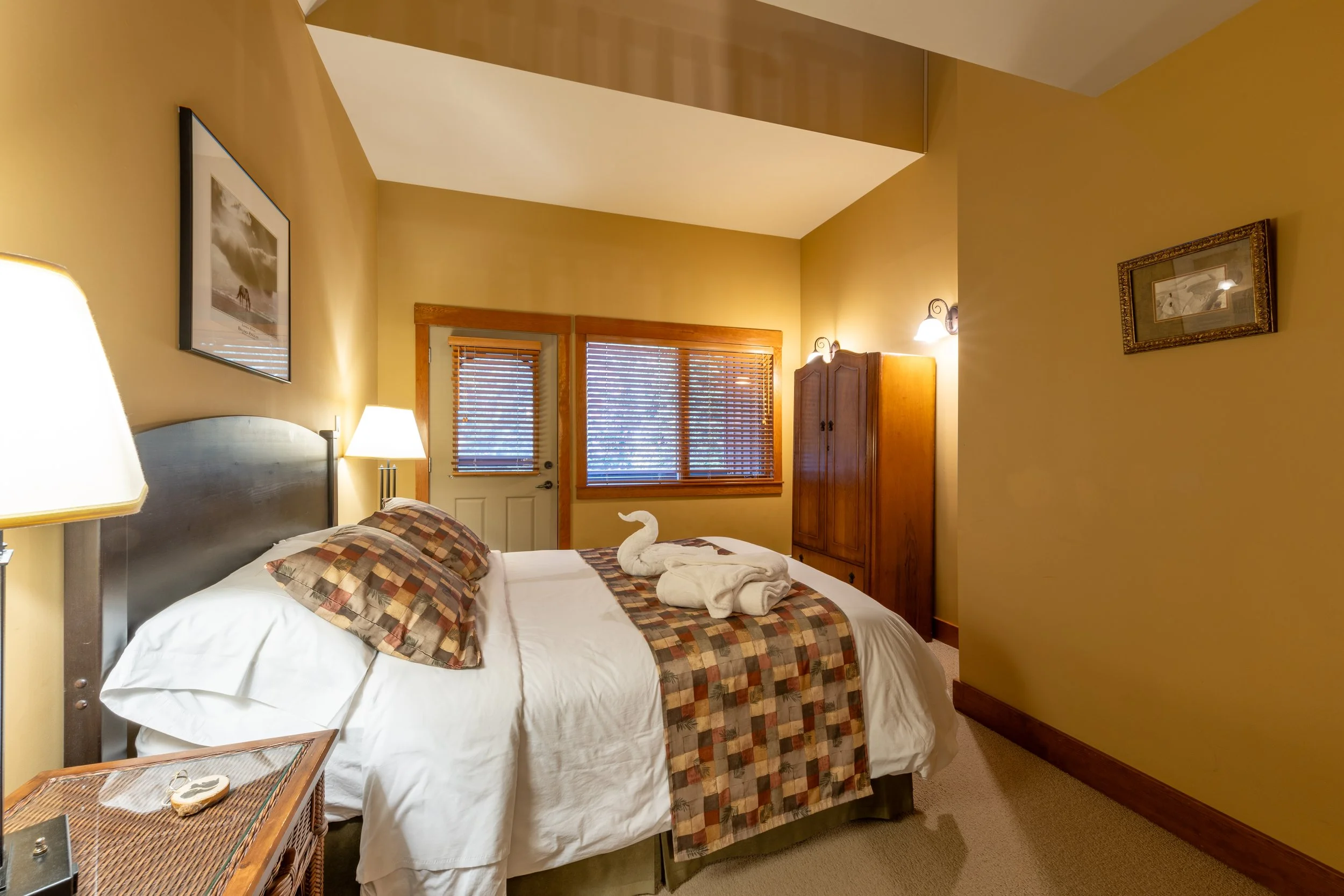 A cozy bedroom with a bed covered in white linens and plaid bedding. There are two folded towels and a towel shaped like a swan on the bed. The room has tan walls, wooden window frames, and blinds, with warm lighting from wall-mounted lamps. Artwork decorates the walls, and a wooden wardrobe is in the corner.