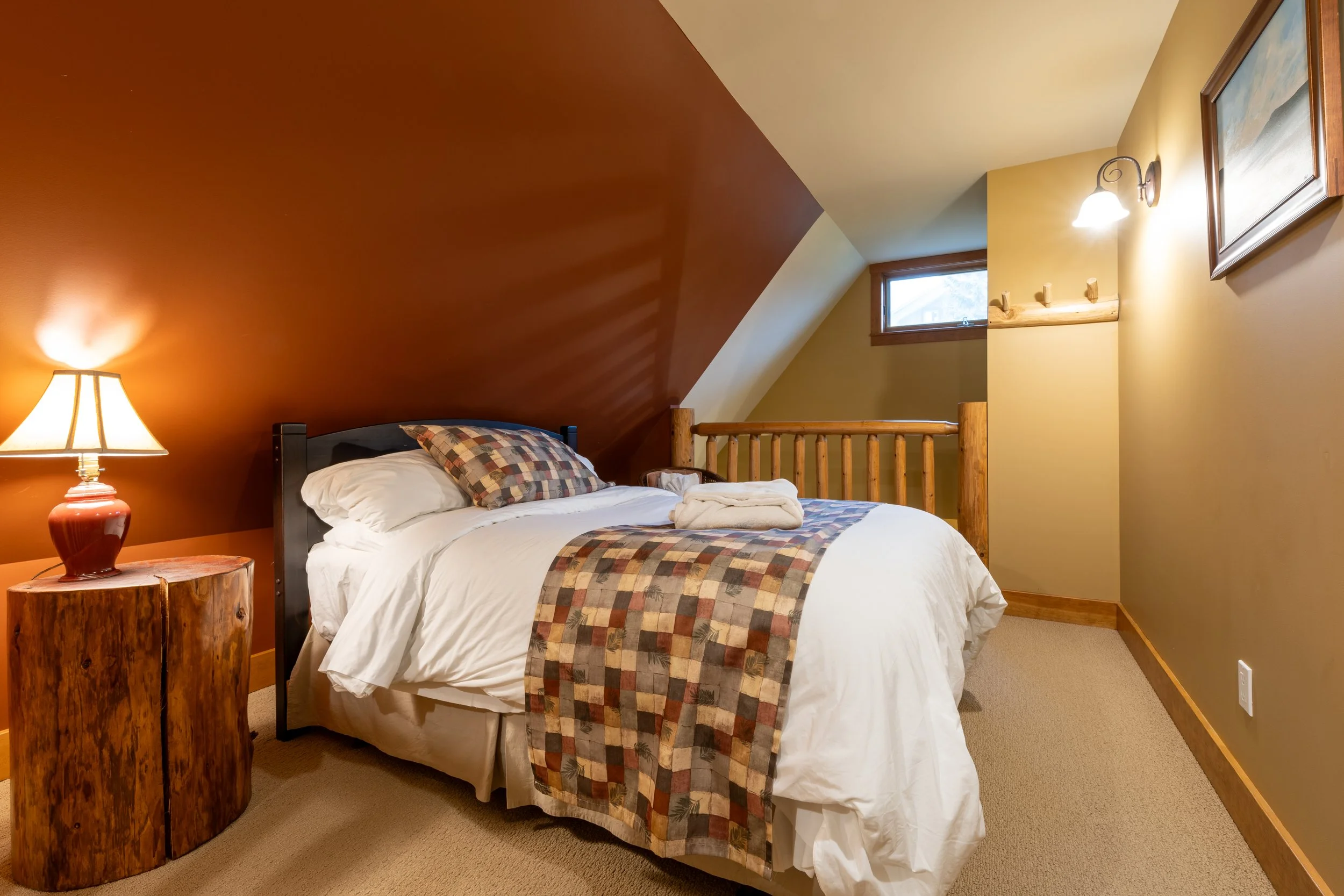 A cozy bedroom with a bed, patterned bedding, a rustic wooden nightstand with a lamp, and a yellow wall with a framed picture and a wall sconce. There are small windows near the ceiling letting in natural light.
