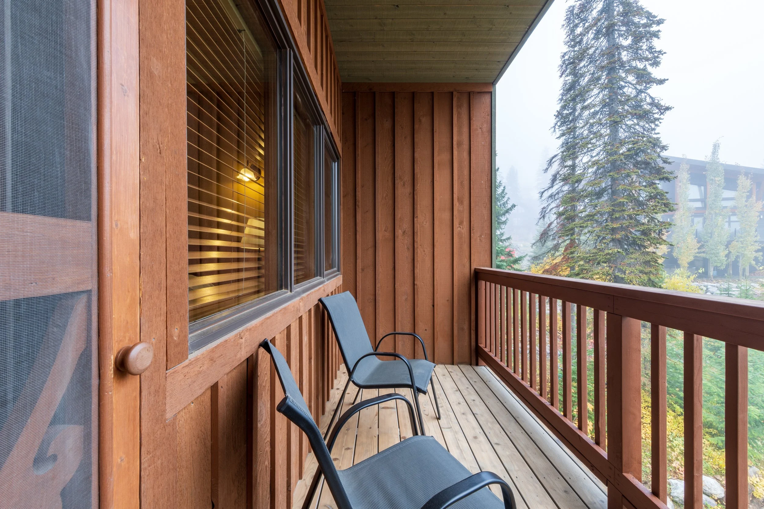 A balcony with wooden walls and floor, three black chairs, and a view of tall trees and foggy sky in a forested area.