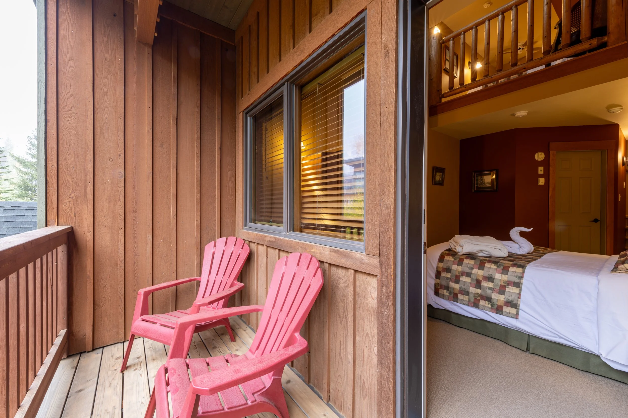 A balcony with pink Adirondack chairs, wooden railing, and windows looking into a bedroom with a bed, folded towels shaped like a swan, and wooden interior.