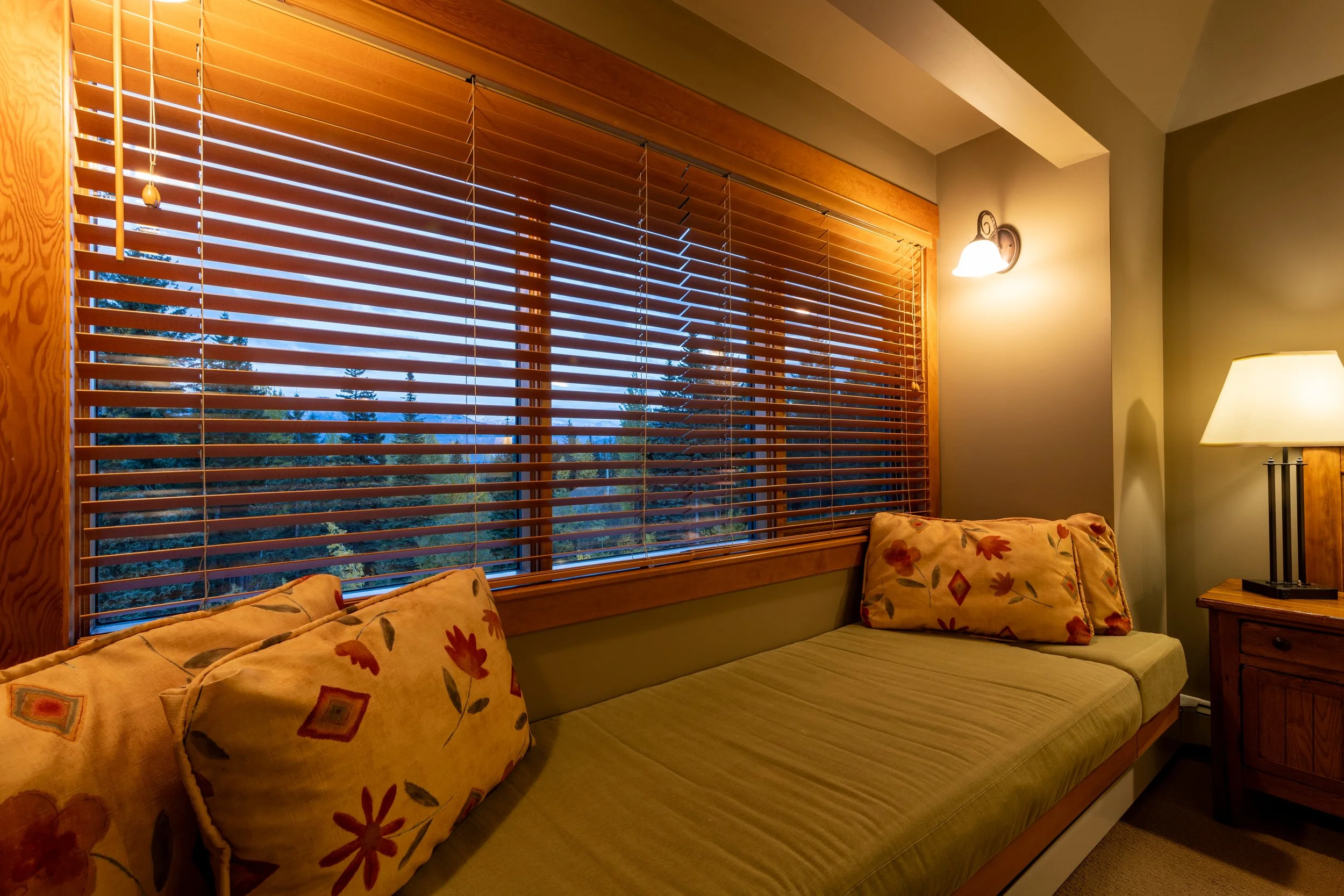 Bedroom area with large window featuring wooden blinds, a sofa with floral-patterned pillows, a side table with a lamp, and a wall-mounted light fixture.