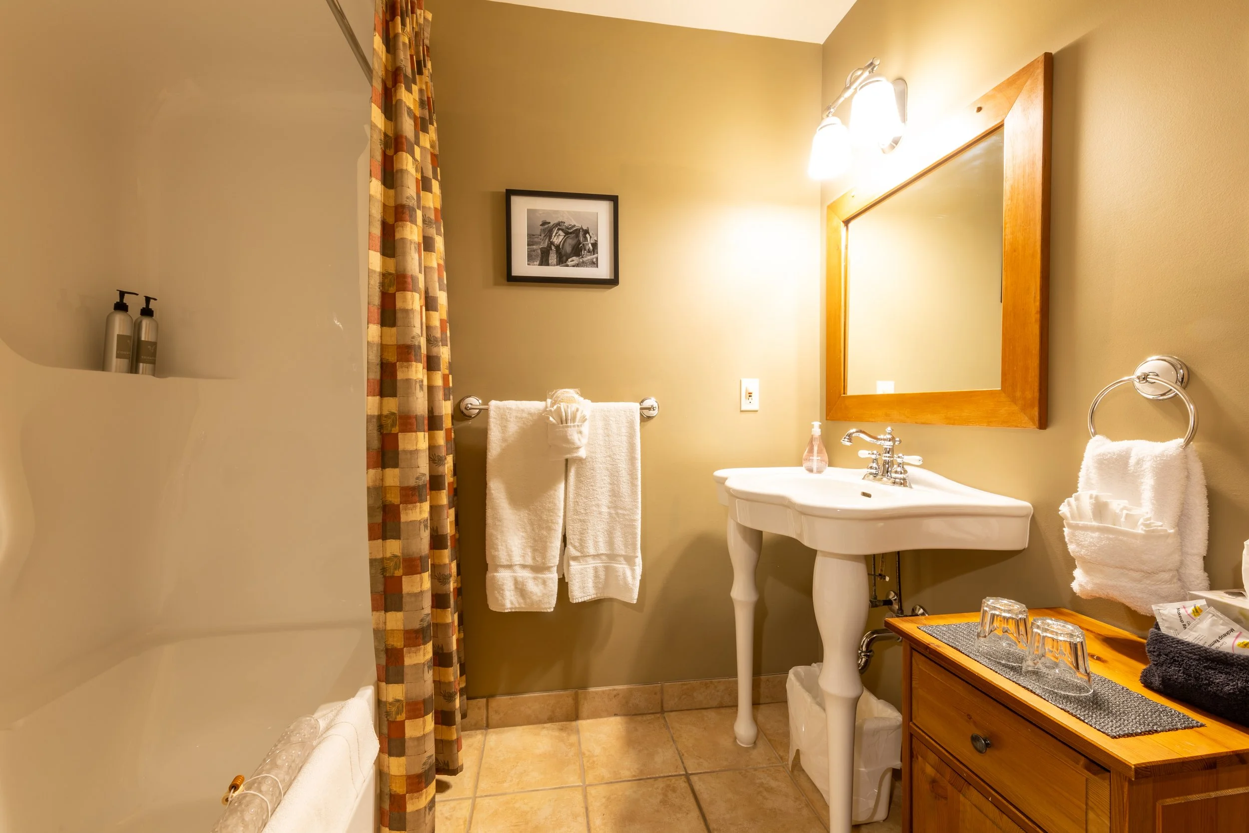 A cozy bathroom with a small white pedestal sink, a wicker storage unit, and a mirror framed in wood. The bathroom features beige walls, a black-and-white horse photograph on the wall, a towel rack with white towels, a decorative curtain with a colorful checkered pattern, and a corner shower with a wall niche holding bottles. The countertop holds a hand soap dispenser, glasses, and some toiletries.