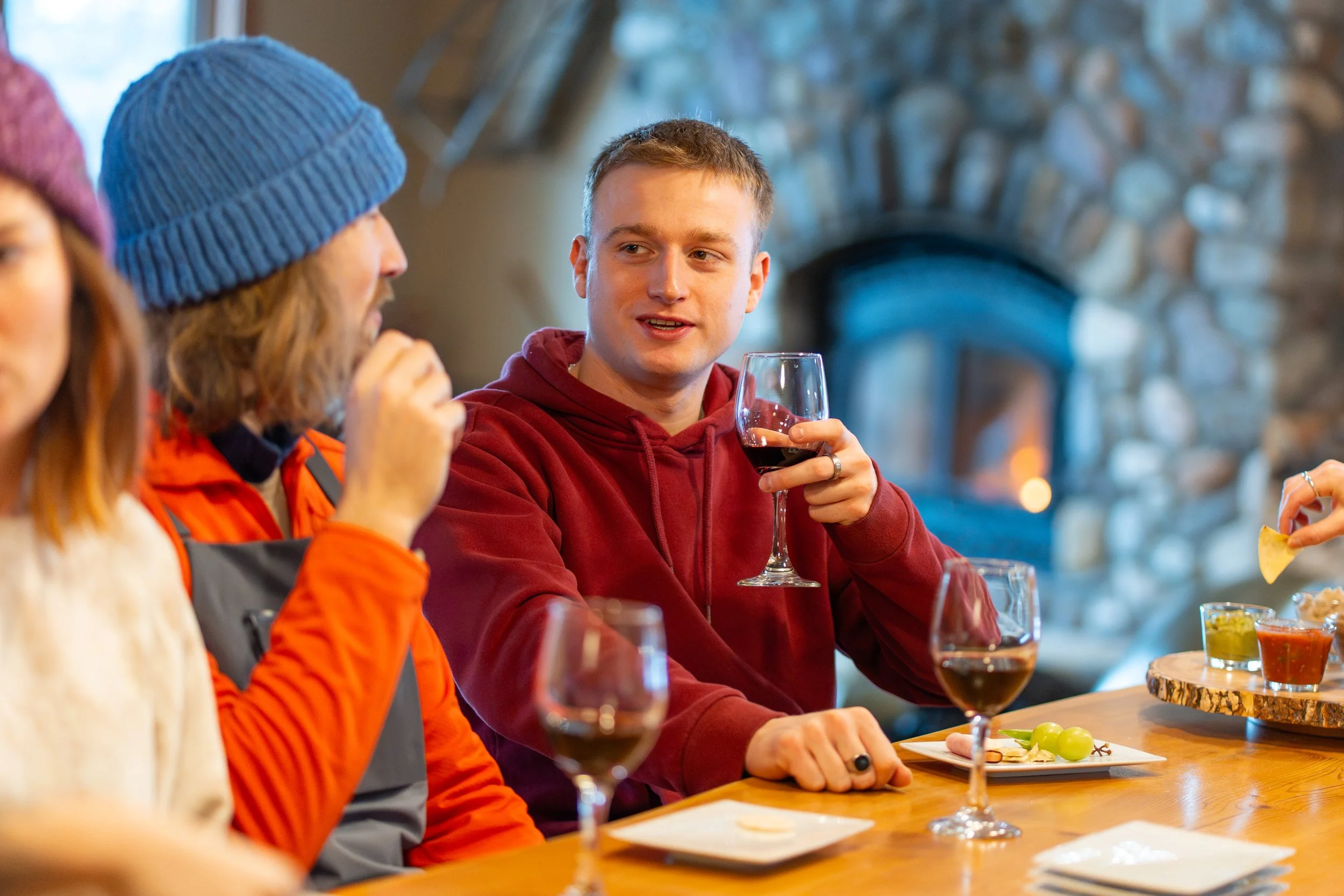 People sitting at a table enjoying drinks and food, with a stone fireplace in the background.