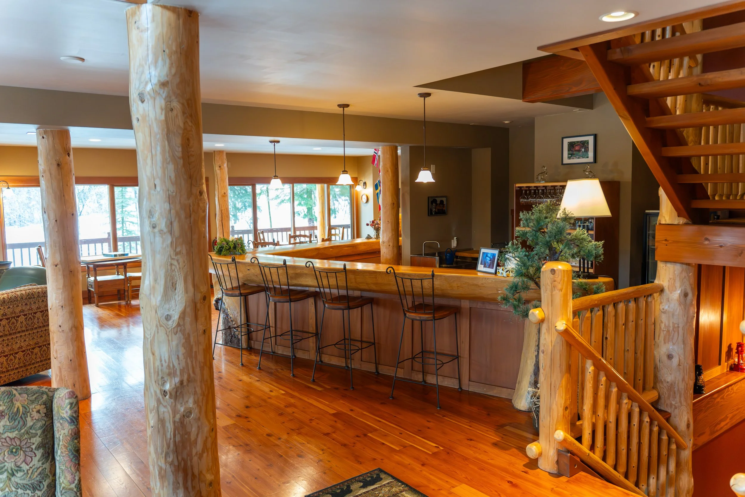 Interior of a rustic lodge with wooden logs and stairs, a bar area with bar stools, and large windows letting in natural light, decorated with greenery and ornaments.