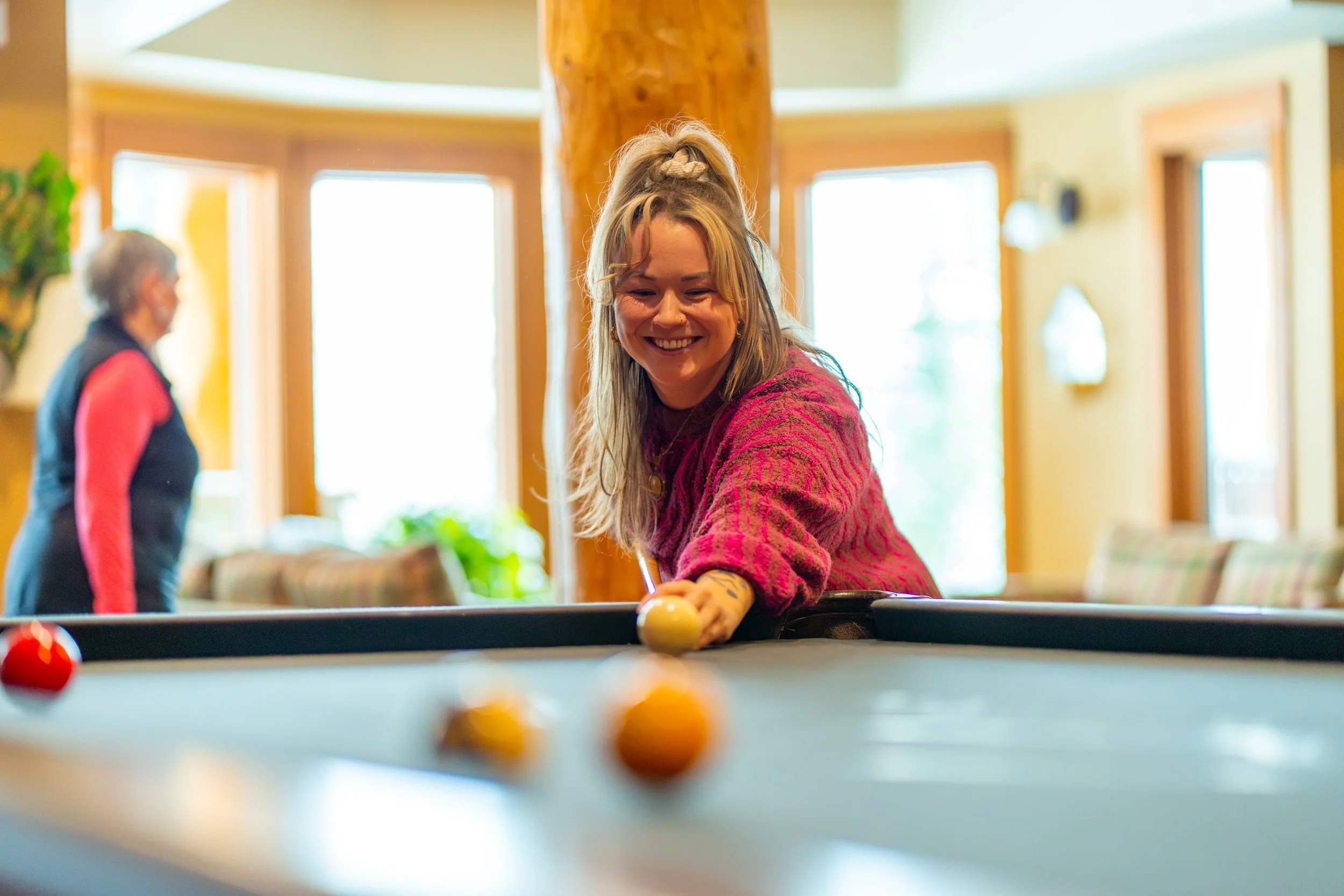 A young woman is playing pool indoors, smiling and leaning over the table to take her shot, with a focus on the balls on the pool table.