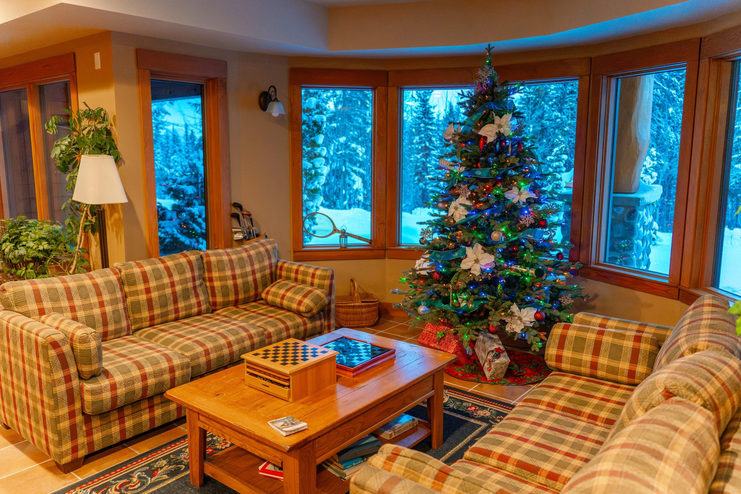A cozy living room decorated for Christmas with a decorated Christmas tree and large windows showing a snowy landscape outside.