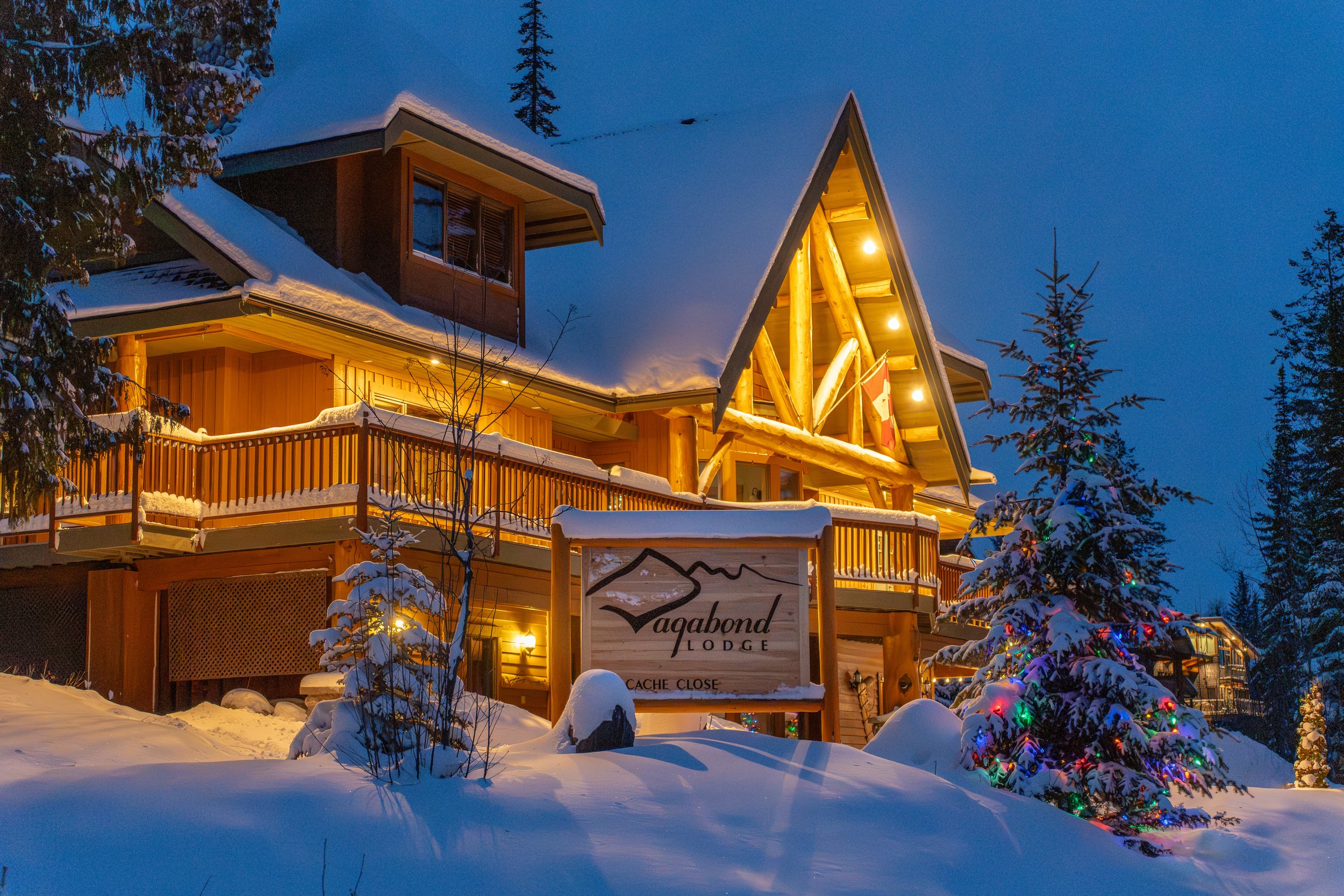 A cozy lodge decorated with Christmas lights during winter snowfall, with snow-covered trees and a blue evening sky in the background.