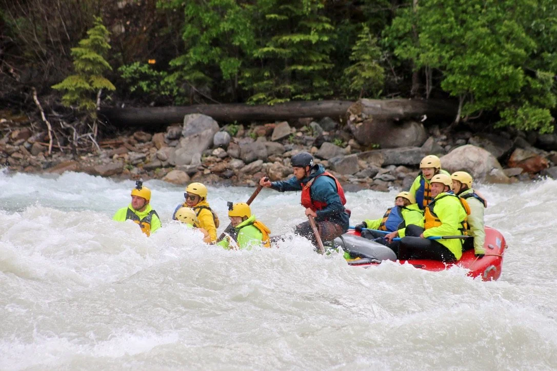 Group of people on a red inflatable raft whitewater rafting in a river, wearing yellow helmets and life jackets, with a guide paddling in front surrounded by lush green trees.