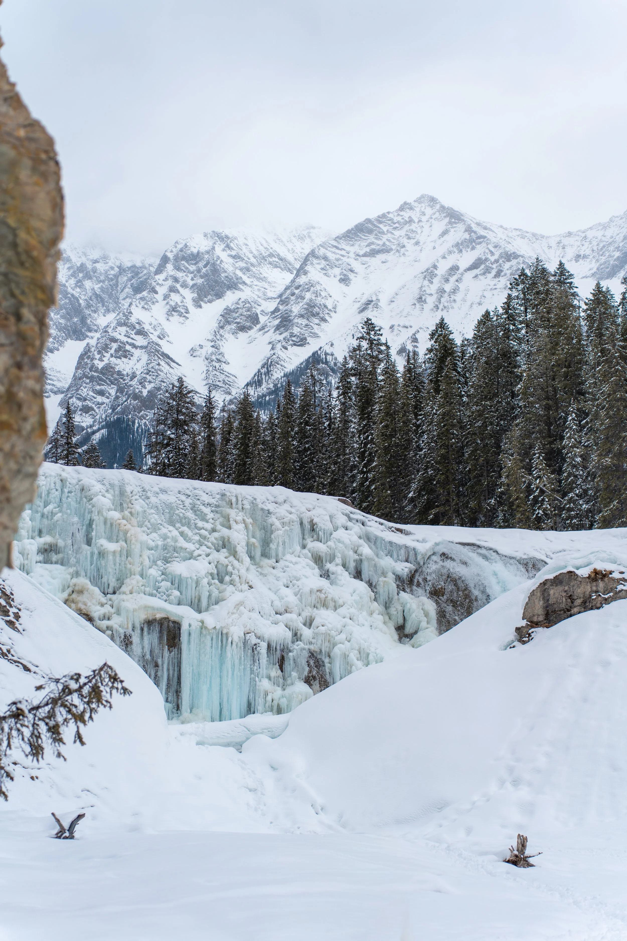 Snow-covered landscape with frozen waterfall in the foreground, pine trees, and tall snow-capped mountains in the background under an overcast sky.