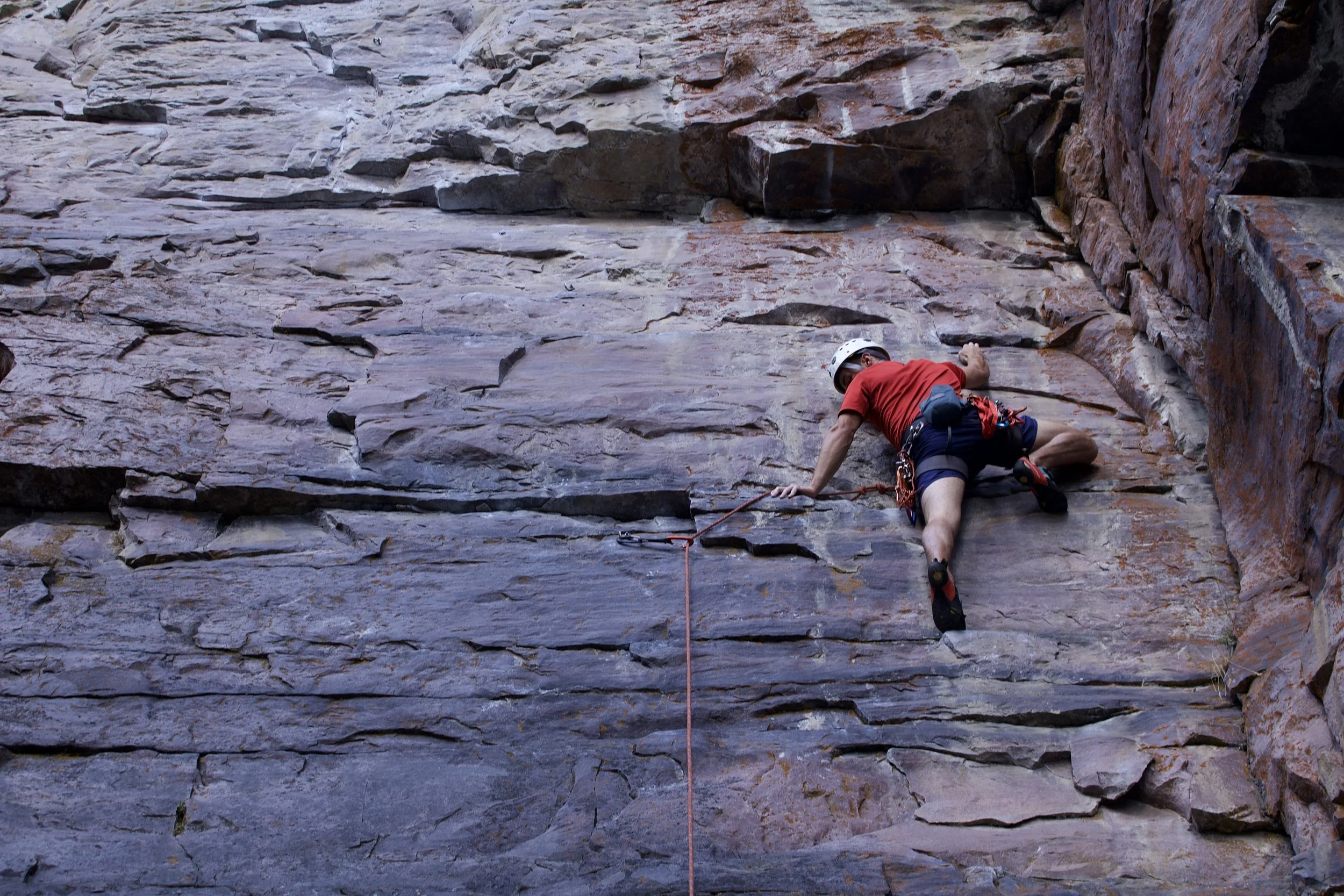 A man wearing a white helmet, red shirt, and shorts rock climbing on a vertical sandstone rock face with climbing gear and a safety rope.