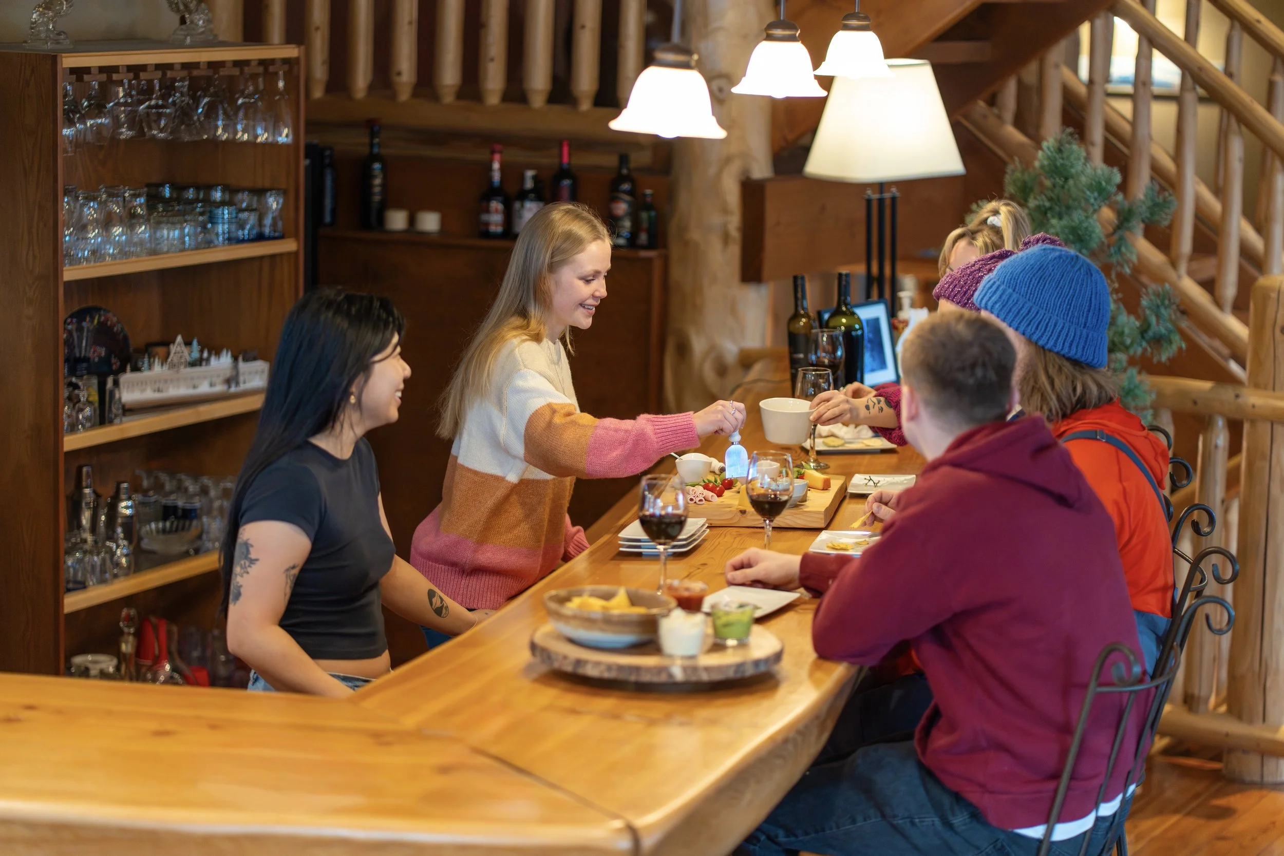 Group of people sitting around a wooden bar area in a cozy, rustic lodge interior with wooden staircase and decor, enjoying food and drinks.