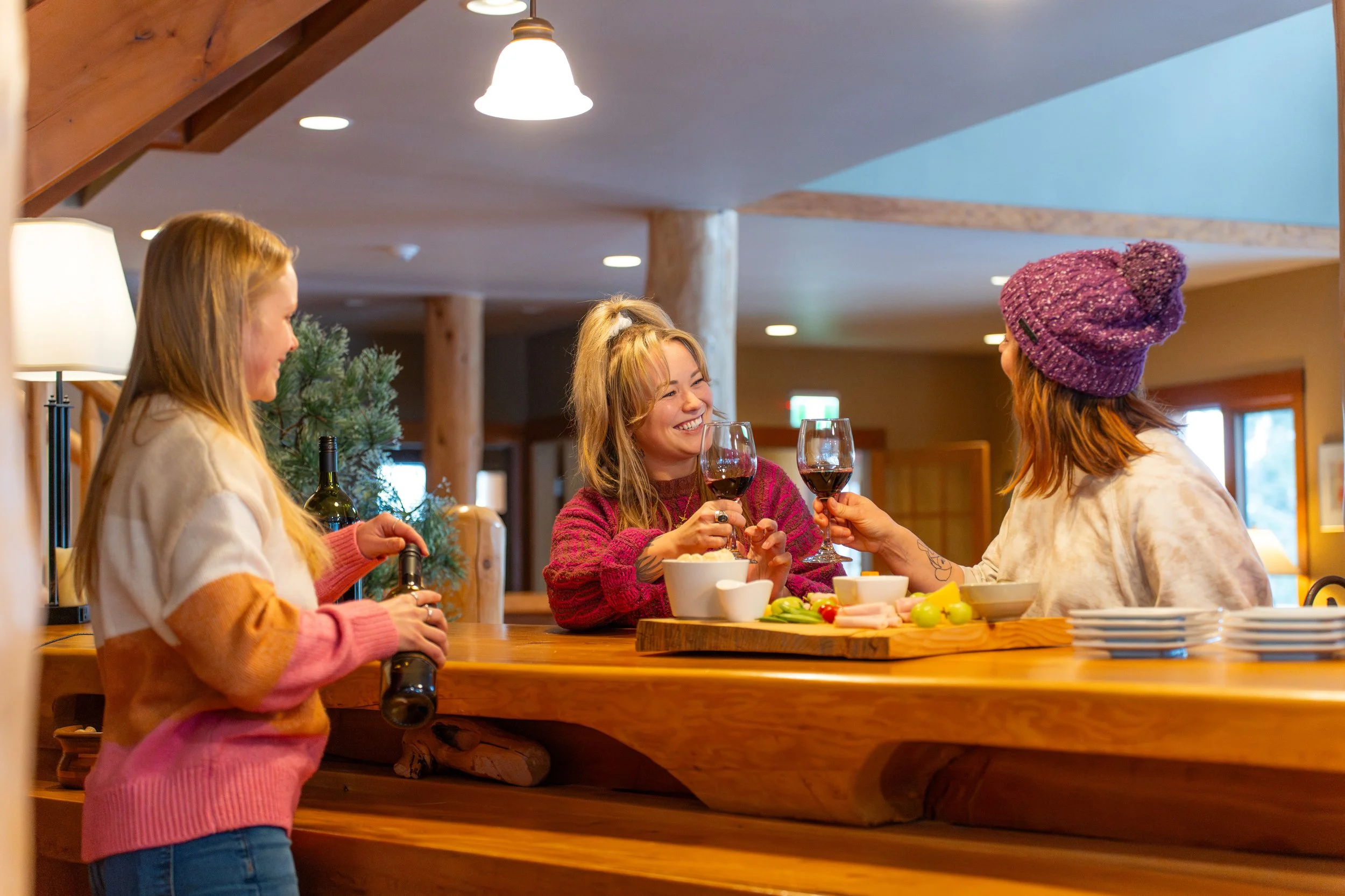 Three women are enjoying a social gathering in a cozy kitchen or dining area, holding glasses of red wine and smiling at each other, with a variety of food on the table.