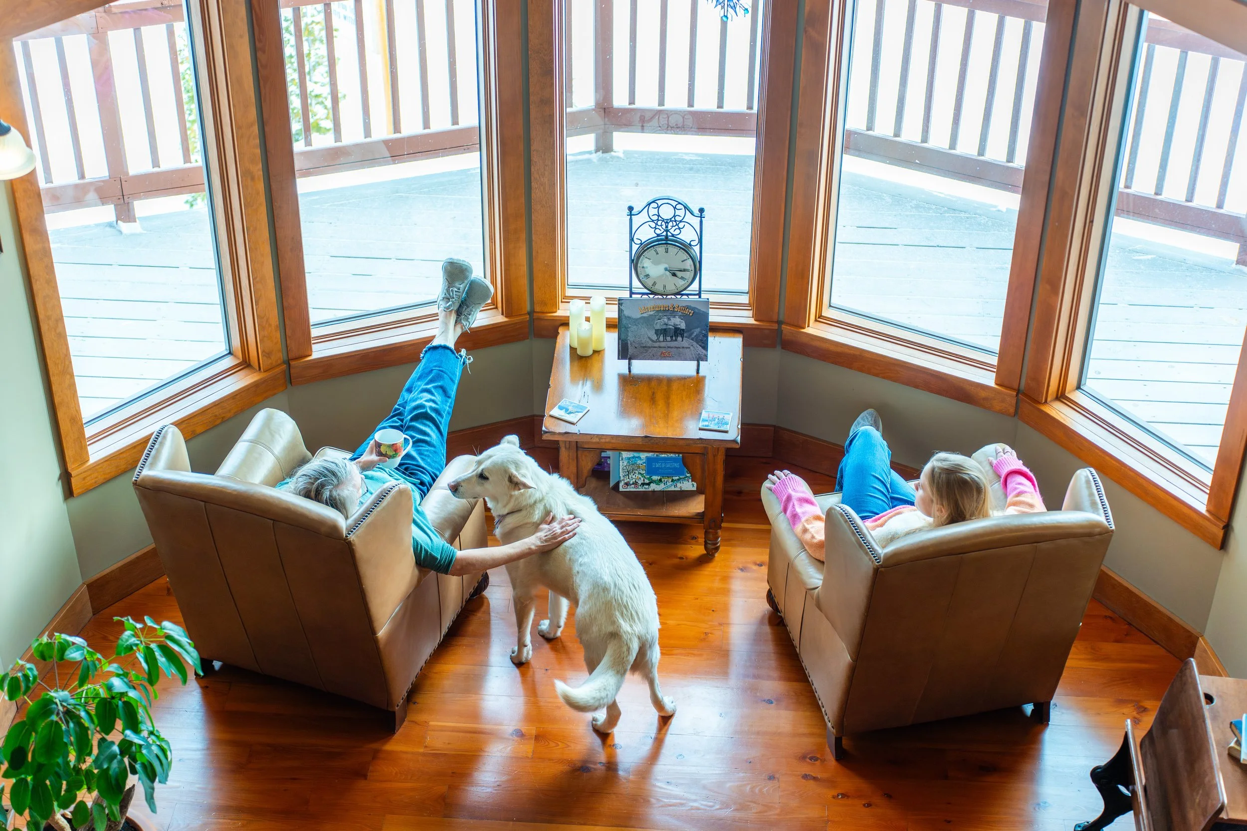 People and dogs sitting on couches in a living room with large bay windows, a clock, candles, and a table with magazines.