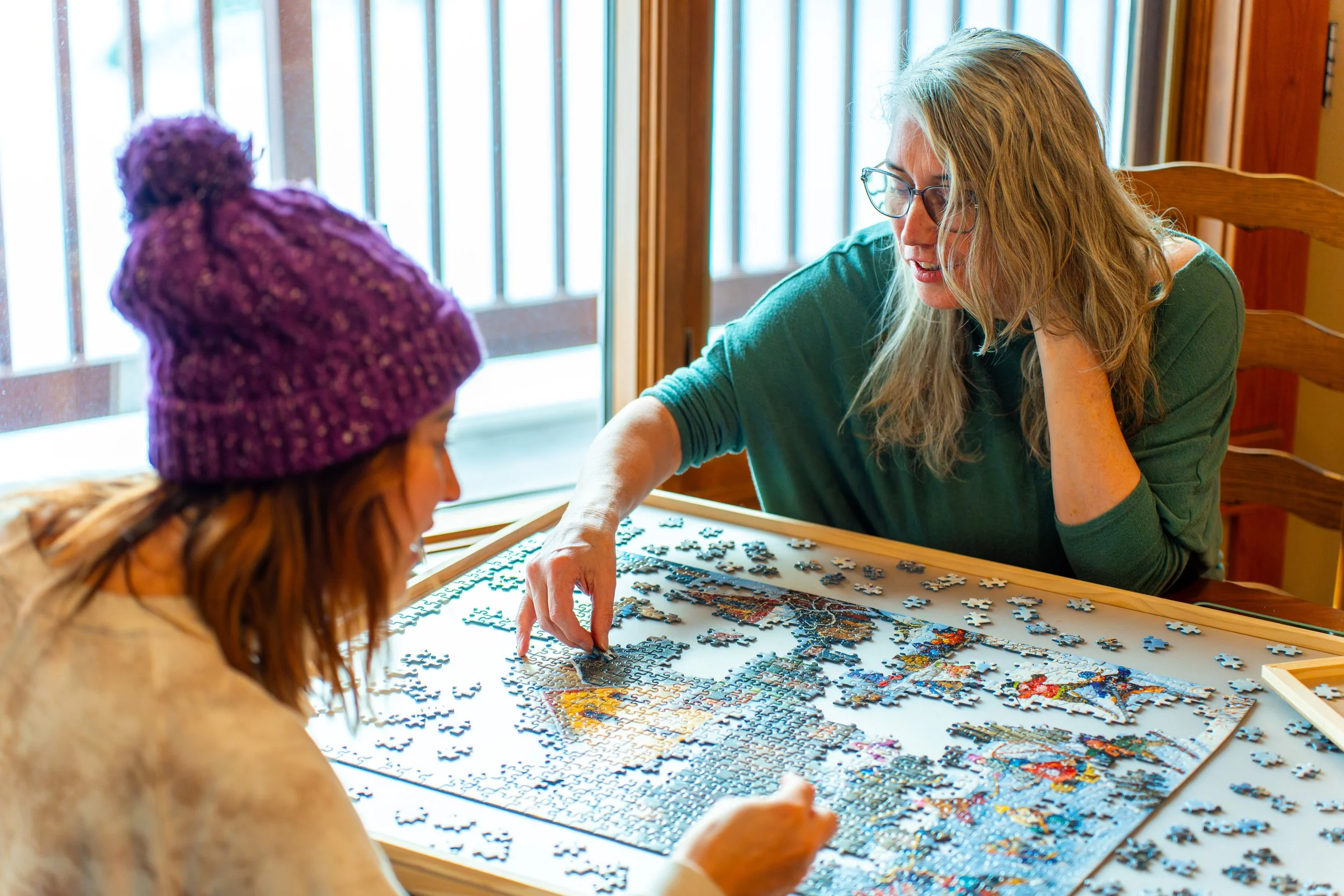 Two women working on a large jigsaw puzzle at a table near a window.