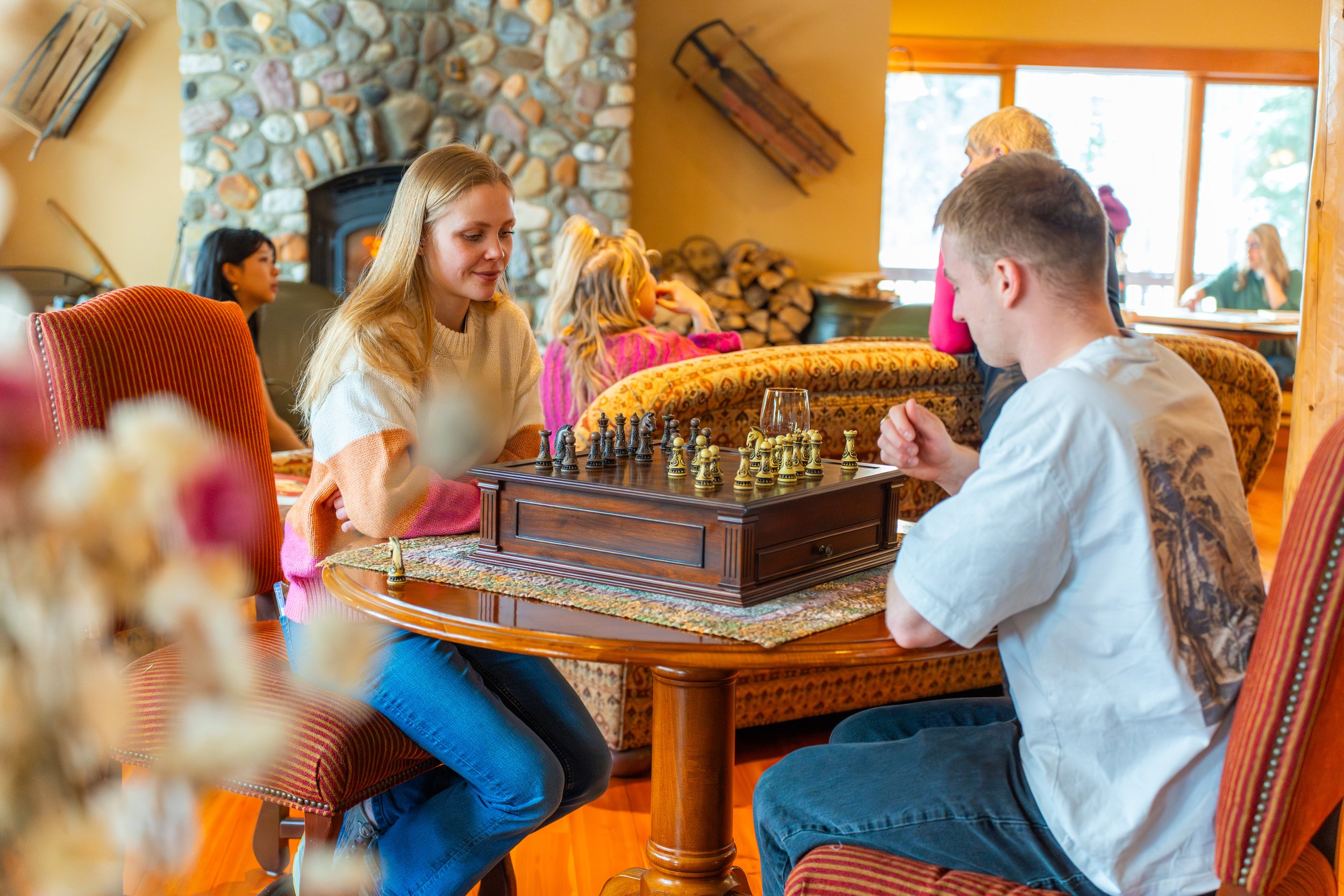 Two young people, a girl with long blonde hair and a boy with short hair, are playing chess at a round wooden table inside a cozy room with a stone fireplace, wood-paneled walls, and large windows showing trees outside. There are other people in the 