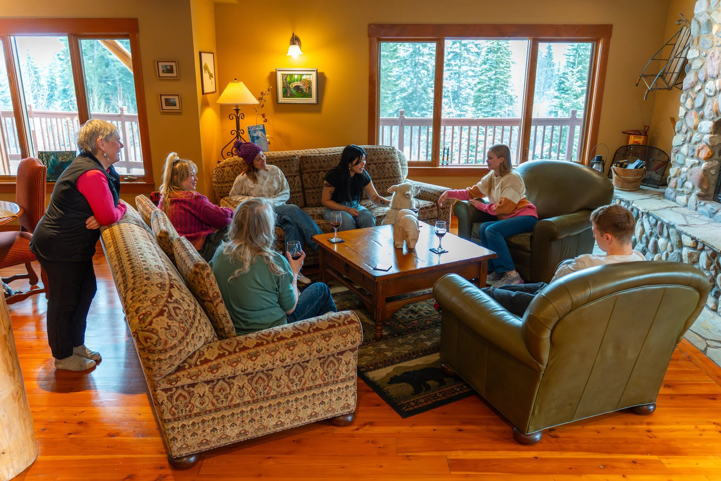 A group of people, mostly women and children, gathered inside a cozy living room with large windows showing a snowy forest outside. They are sitting and standing around a sofa and a coffee table, engaging in a conversation with a woman standing. The 
