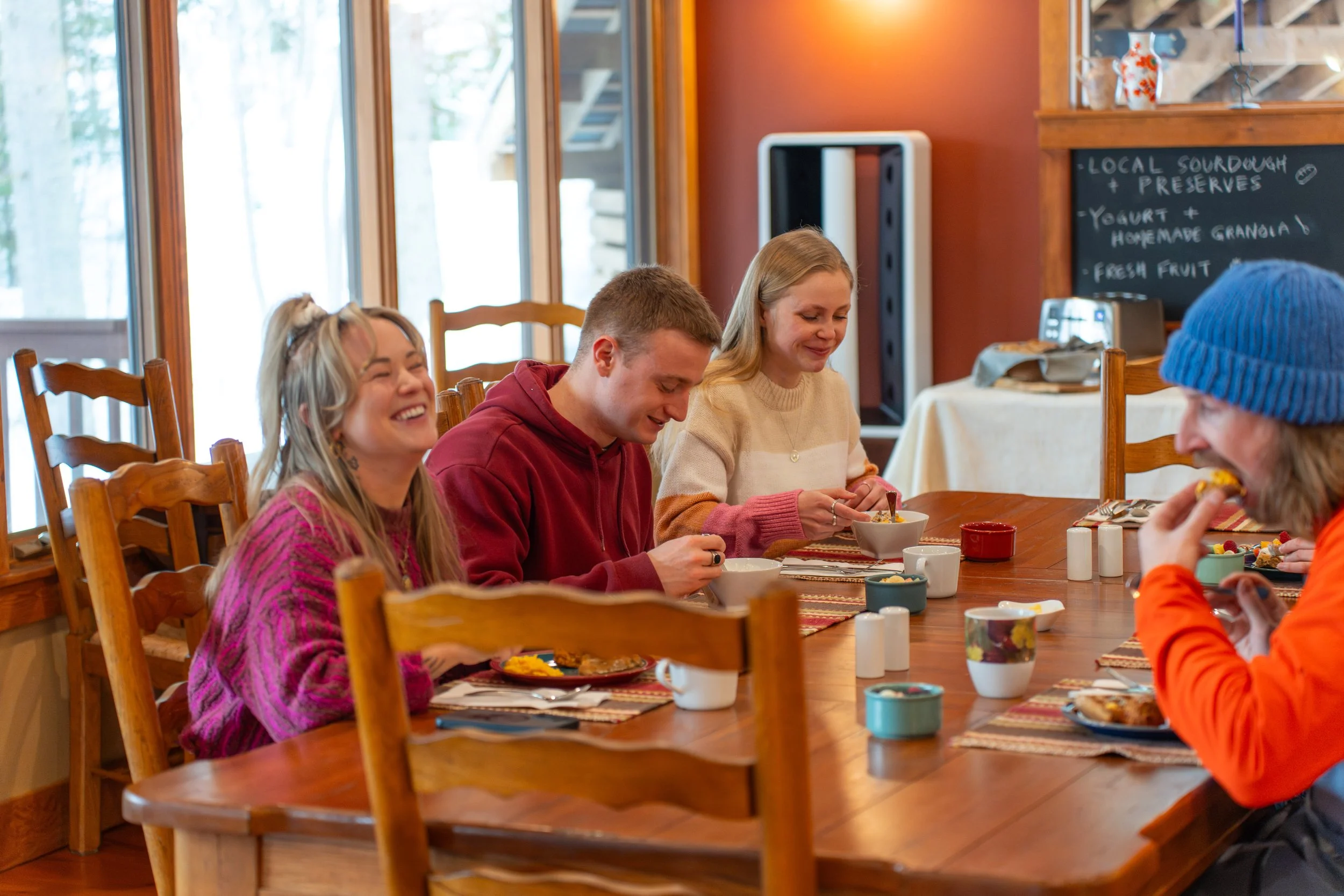 People sitting at a wooden dining table enjoying a meal in a cozy restaurant with large windows and a chalkboard menu in the background.
