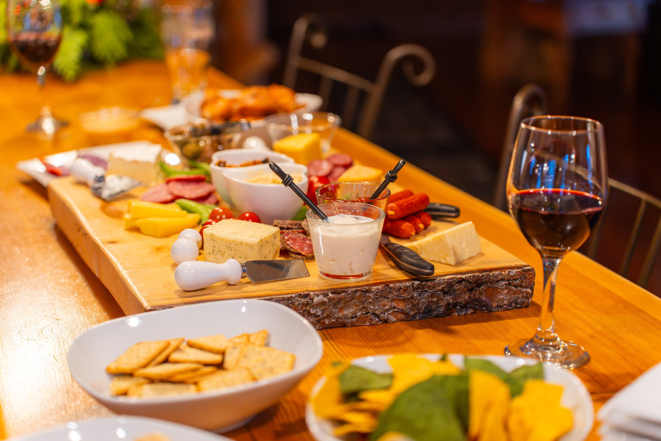 A wooden serving board with assorted cheeses, cured meats, vegetables, dips, and bread on a dining table next to a glass of red wine.