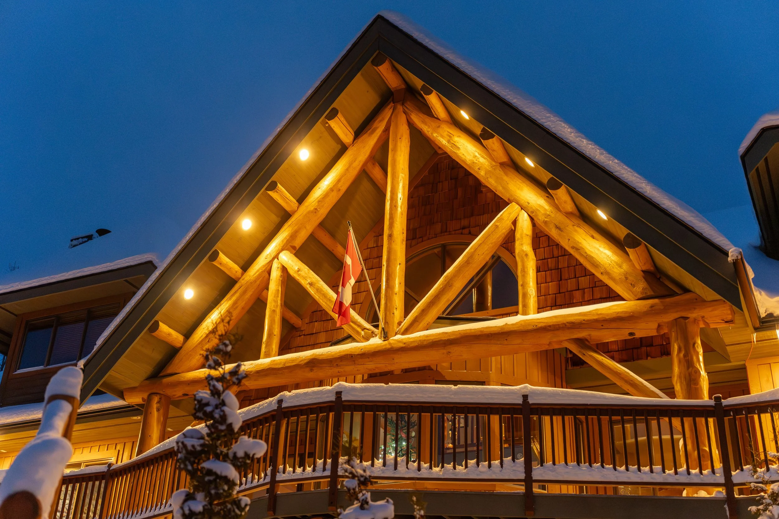 The image shows the front of a house illuminated with warm lights, with large wooden logs supporting the structure's upper part, and snow on the roof and in the surrounding area during dusk or early evening.