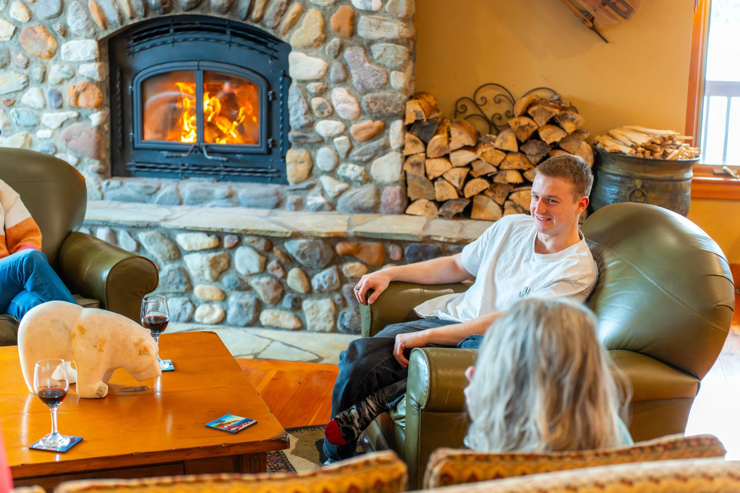 People sitting on a sofa near a fireplace in a cozy living room, with stacked firewood, a coffee table with wine glasses, and a large window in the background.