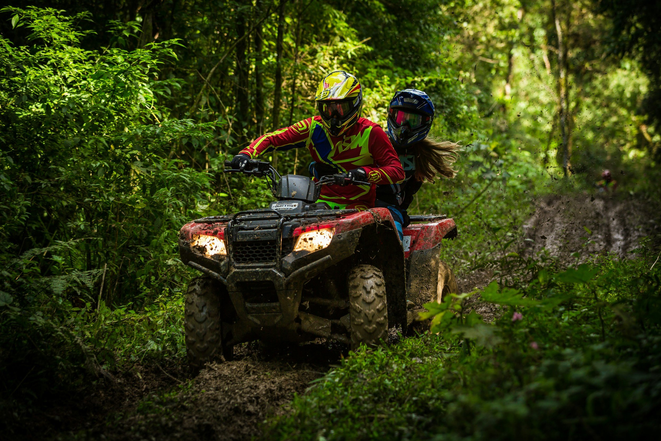 Two people wearing helmets and motocross gear riding an all-terrain vehicle through a dense, green forest trail with mud splashing.