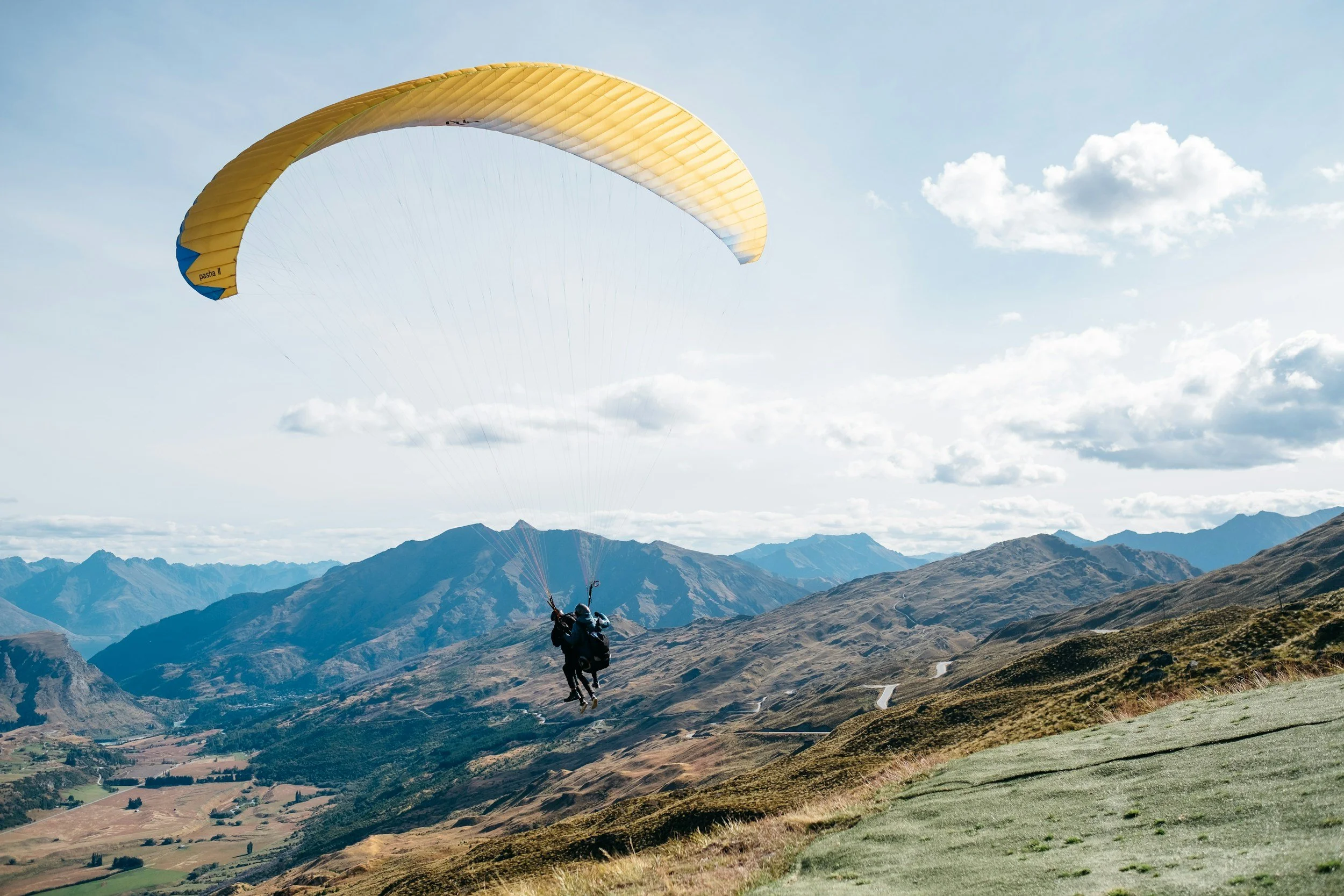 A person parachuting over a mountainous landscape with rolling hills and distant peaks under a partly cloudy sky.