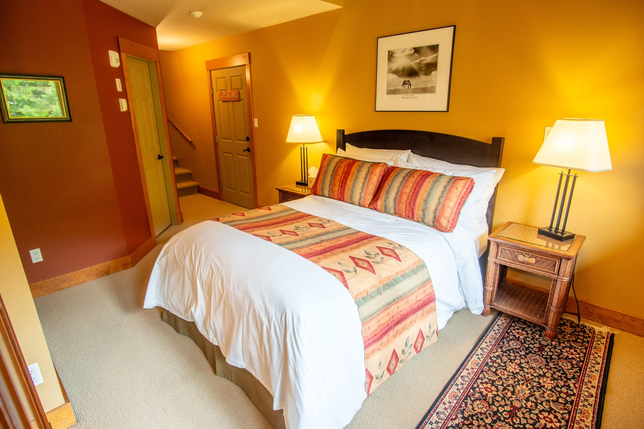 A cozy bedroom with a bed, patterned bedding, a rustic wooden nightstand with a lamp, and a yellow wall with a framed picture and a wall sconce. There are small windows near the ceiling letting in natural light.