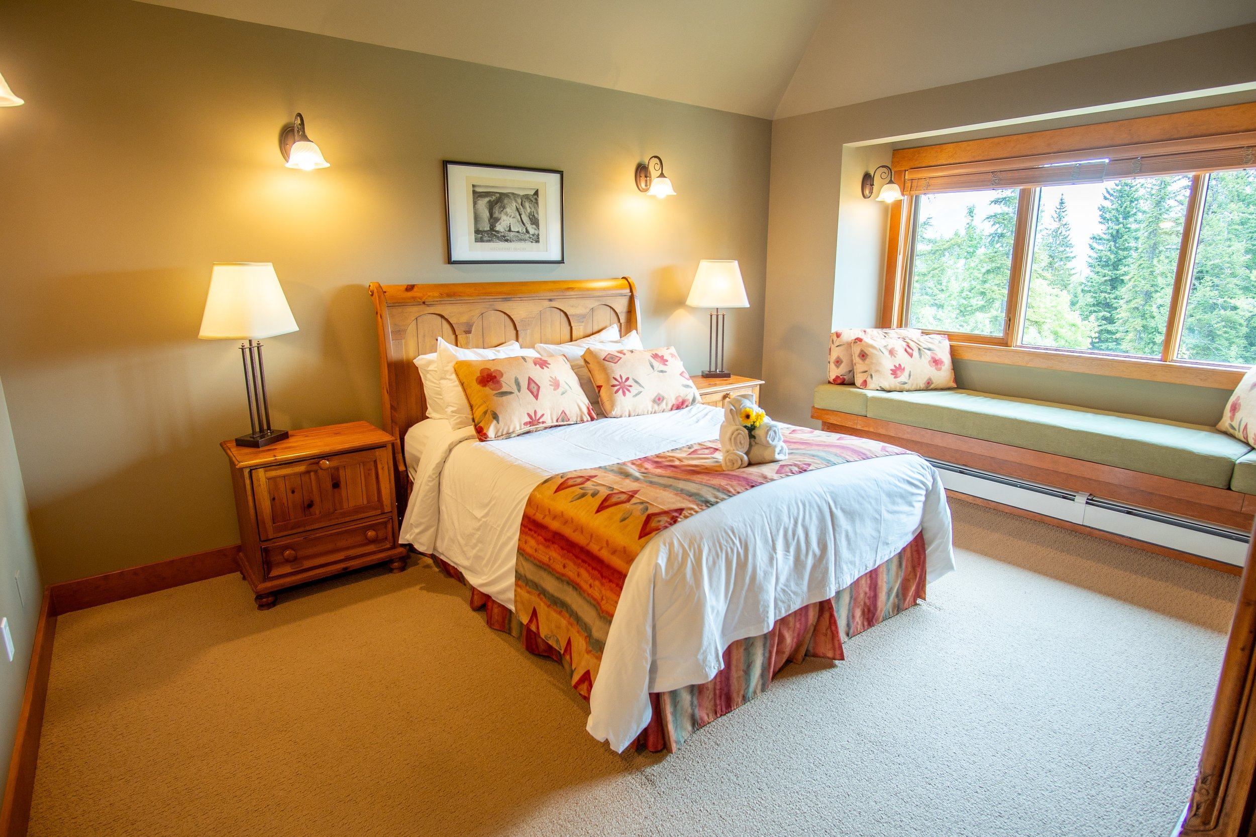 A cozy hotel room with a large bed, wooden furniture, and warm lighting. The bed has a swan towel decoration and colorful pillows. There's a large window with wooden blinds and a built-in bench beneath it. The room features beige walls, a framed picture of a moose, and matching wall sconces.