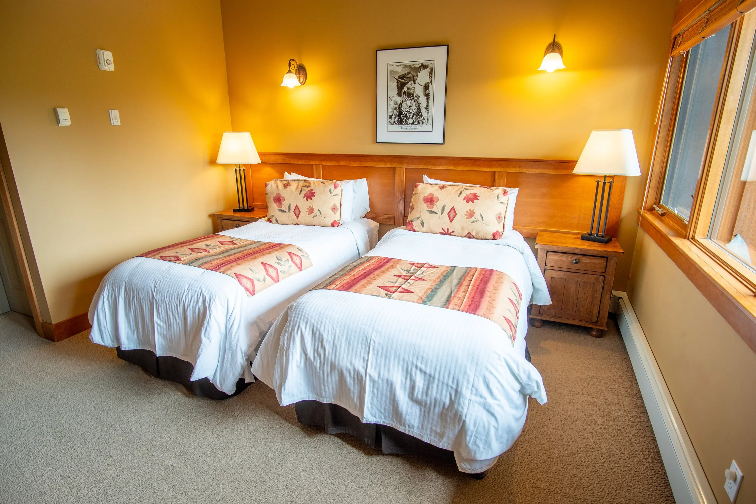A bedroom with two beds, a wooden wardrobe, a window with blinds, and wall-mounted lights.