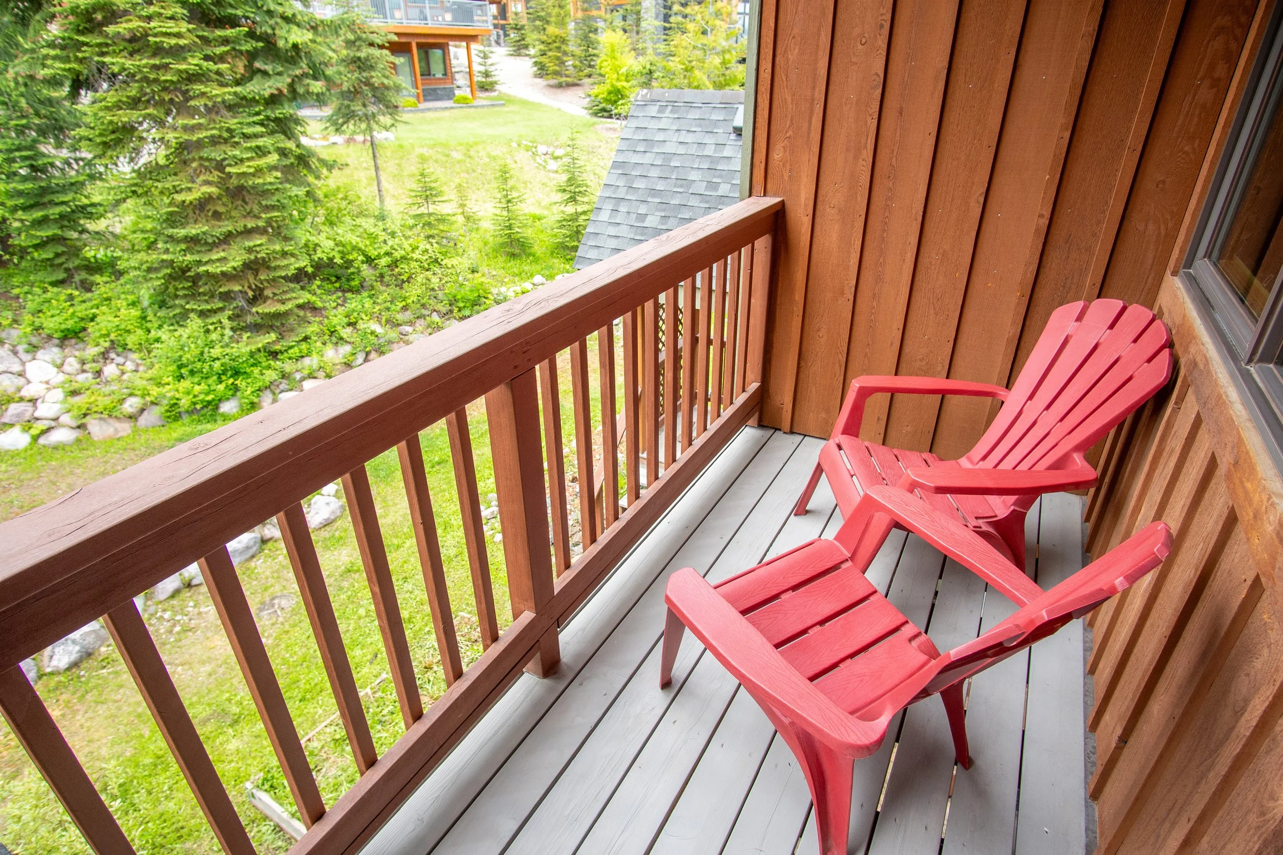 A balcony with pink Adirondack chairs, wooden railing, and windows looking into a bedroom with a bed, folded towels shaped like a swan, and wooden interior.