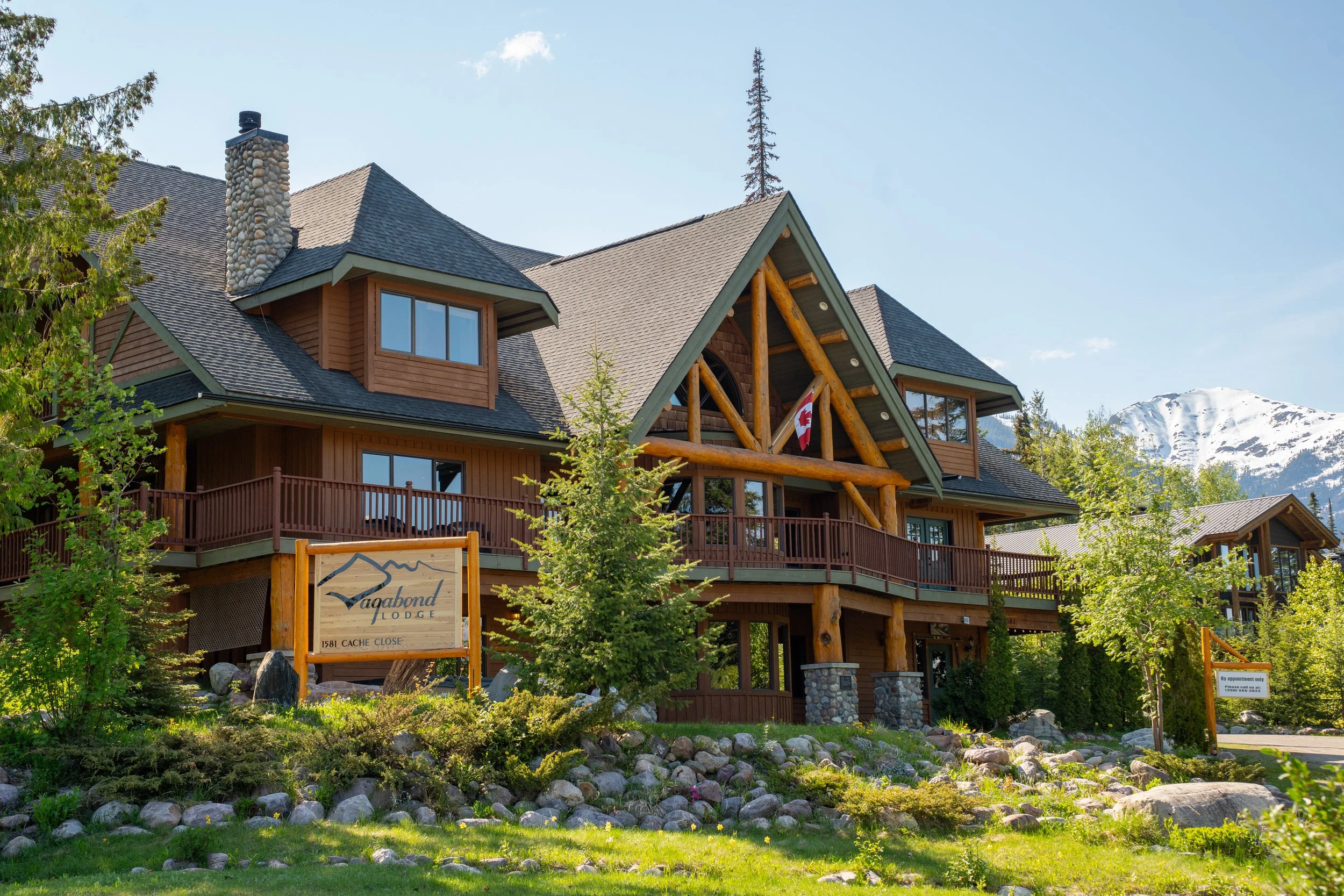 A wooden lodge named Vagabond Lodge with a sign at the front, surrounded by green trees and rocks, with snow-capped mountains in the background, under a clear blue sky.