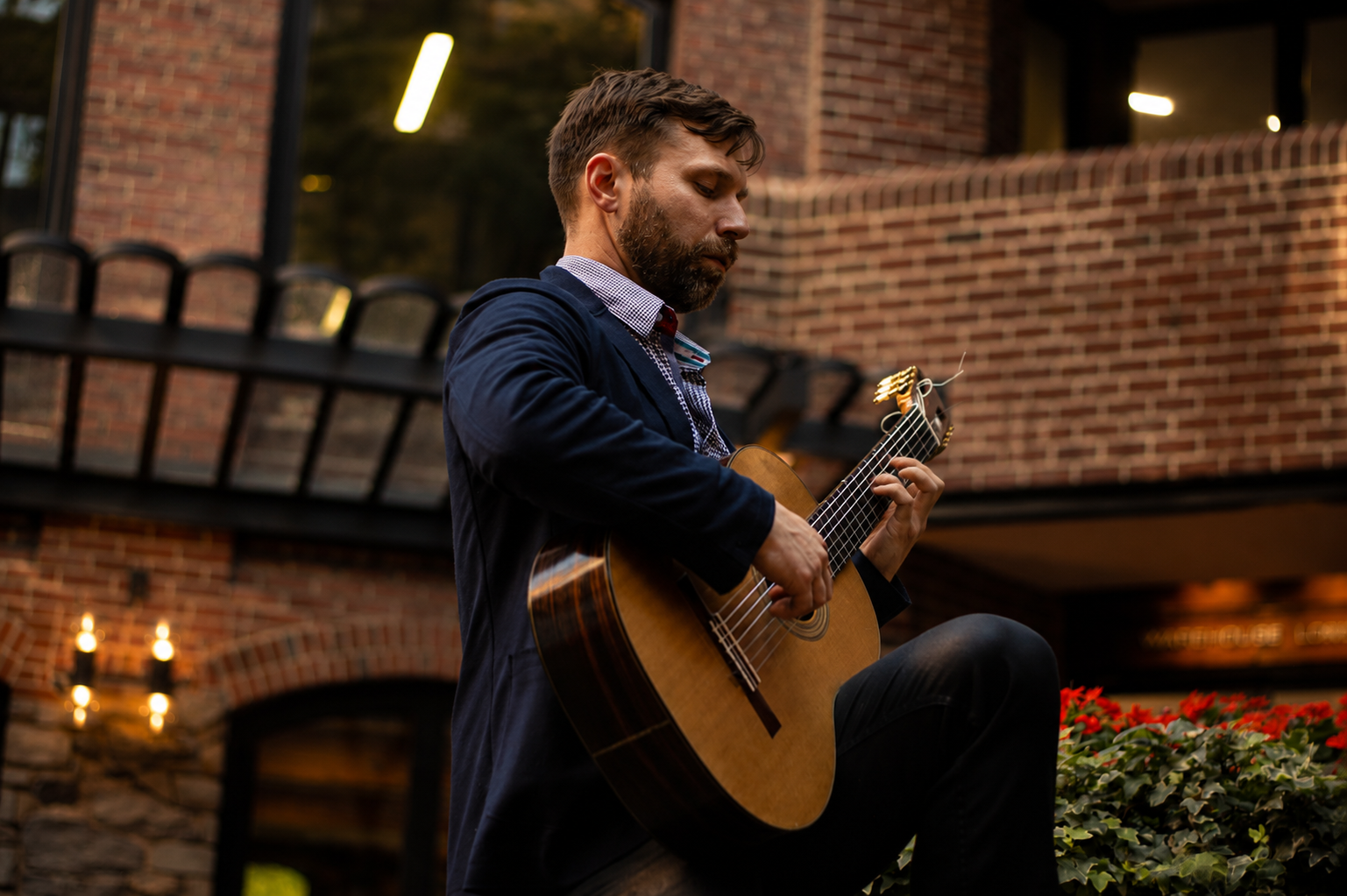 A man playing an acoustic guitar outdoors near a brick building during the evening.