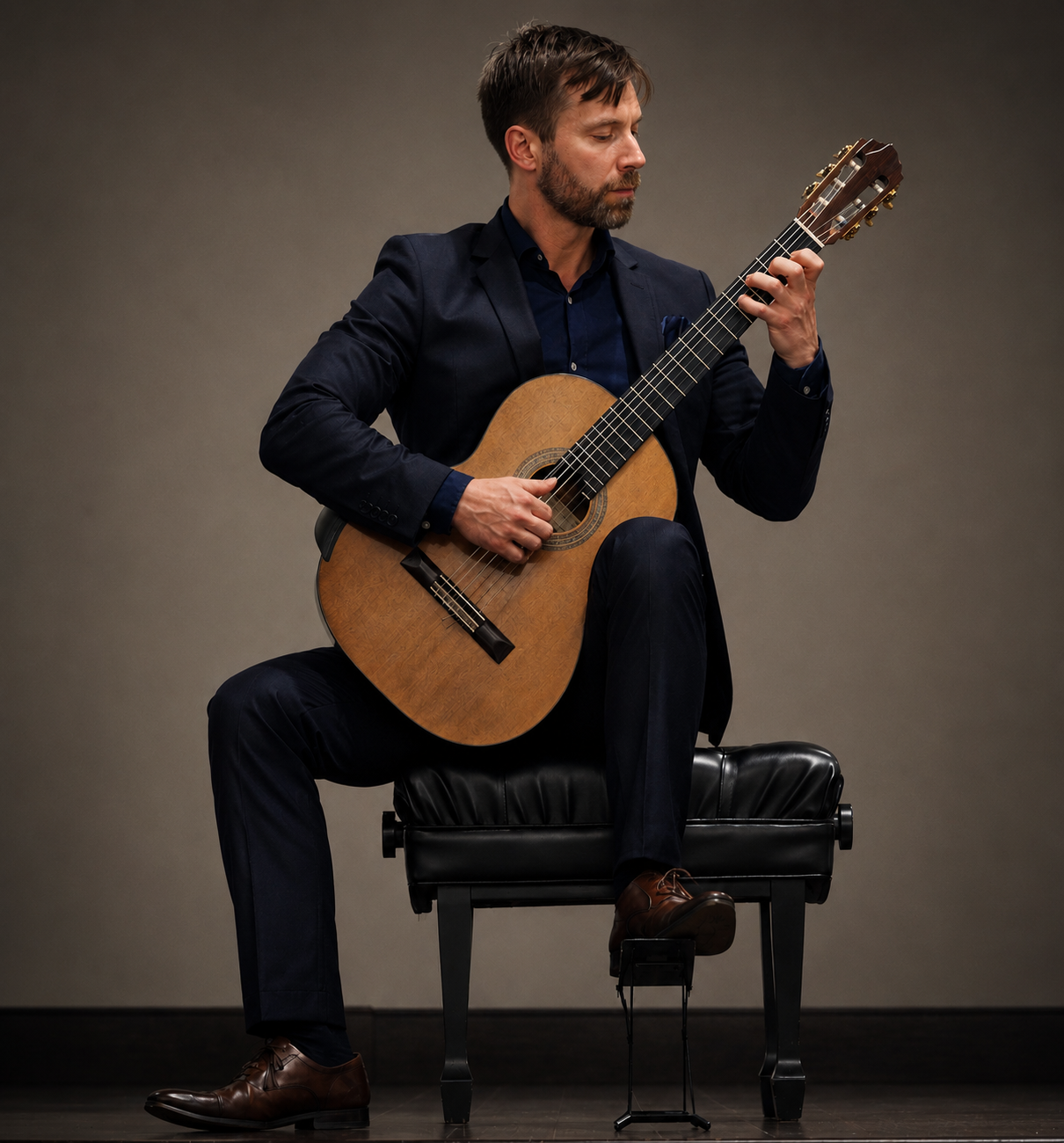 A man in a dark suit sitting on a piano bench, playing an acoustic guitar against a neutral background.