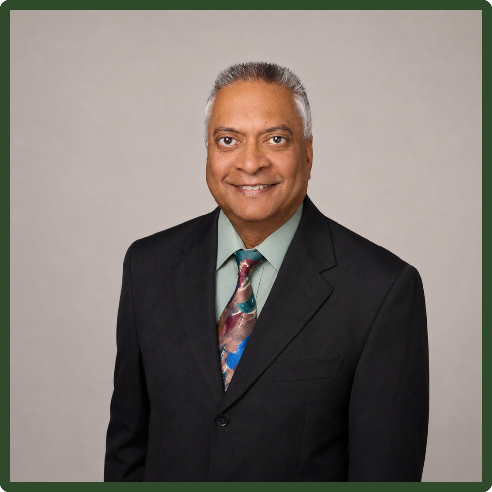 A professional man with gray hair and brown skin wearing a black suit, light green shirt, and a colorful tie, posing against a light gray background.