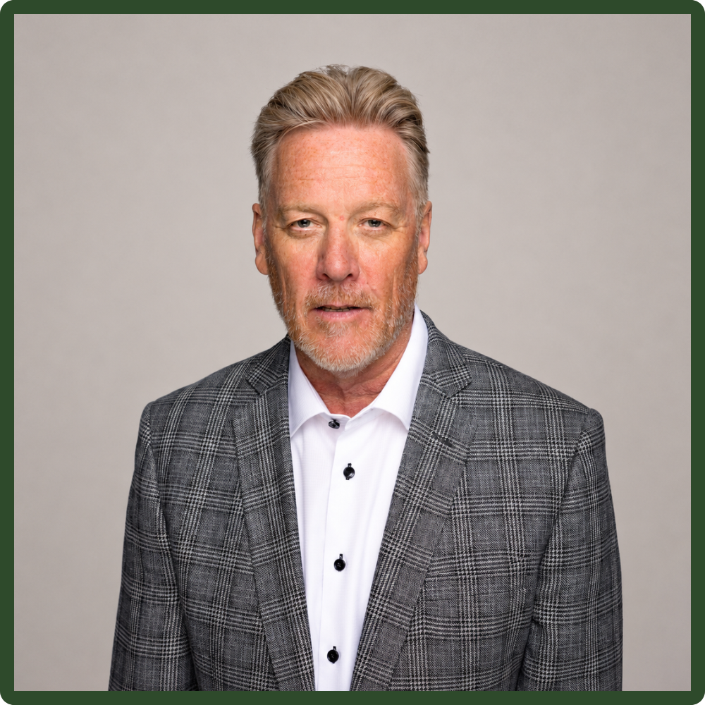 A middle-aged man with gray hair and beard wearing a gray plaid blazer and white shirt standing against a plain gray background.