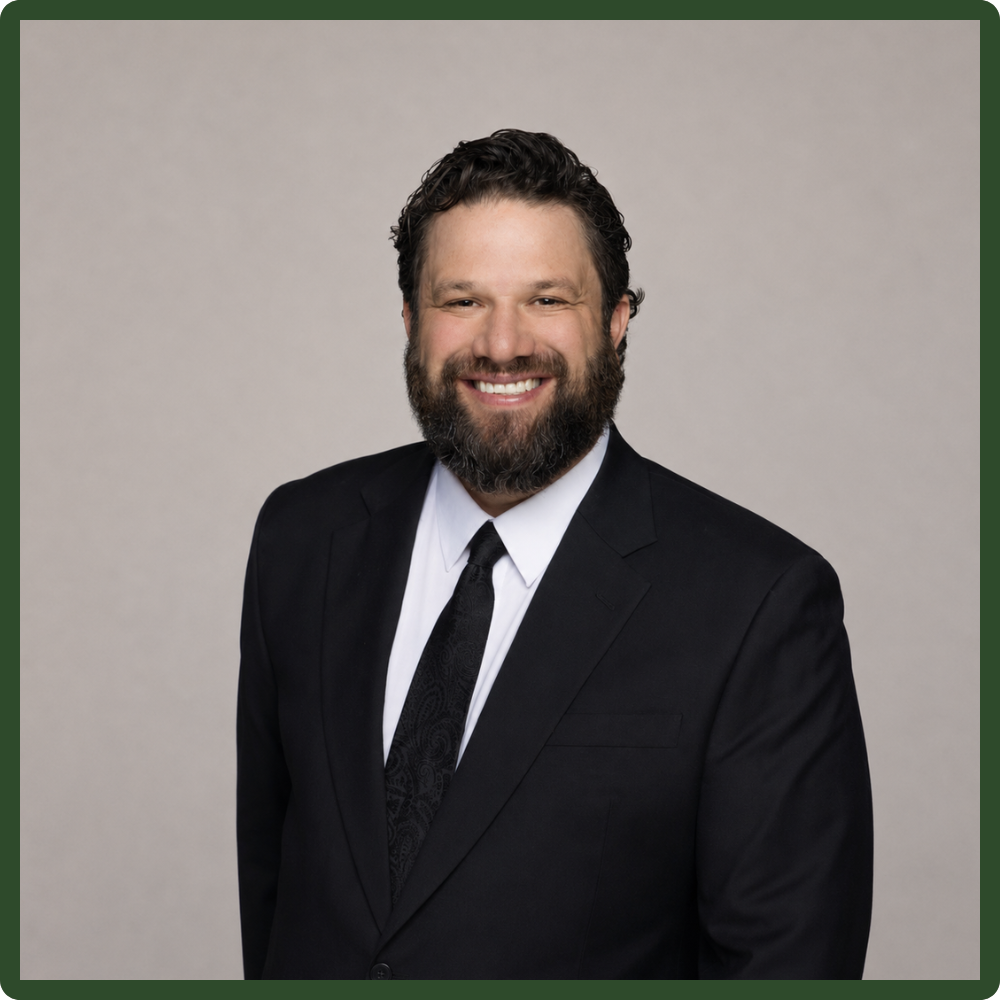 A man with dark curly hair and a beard, wearing a black suit, white shirt, and a black tie, smiling against a plain gray background.
