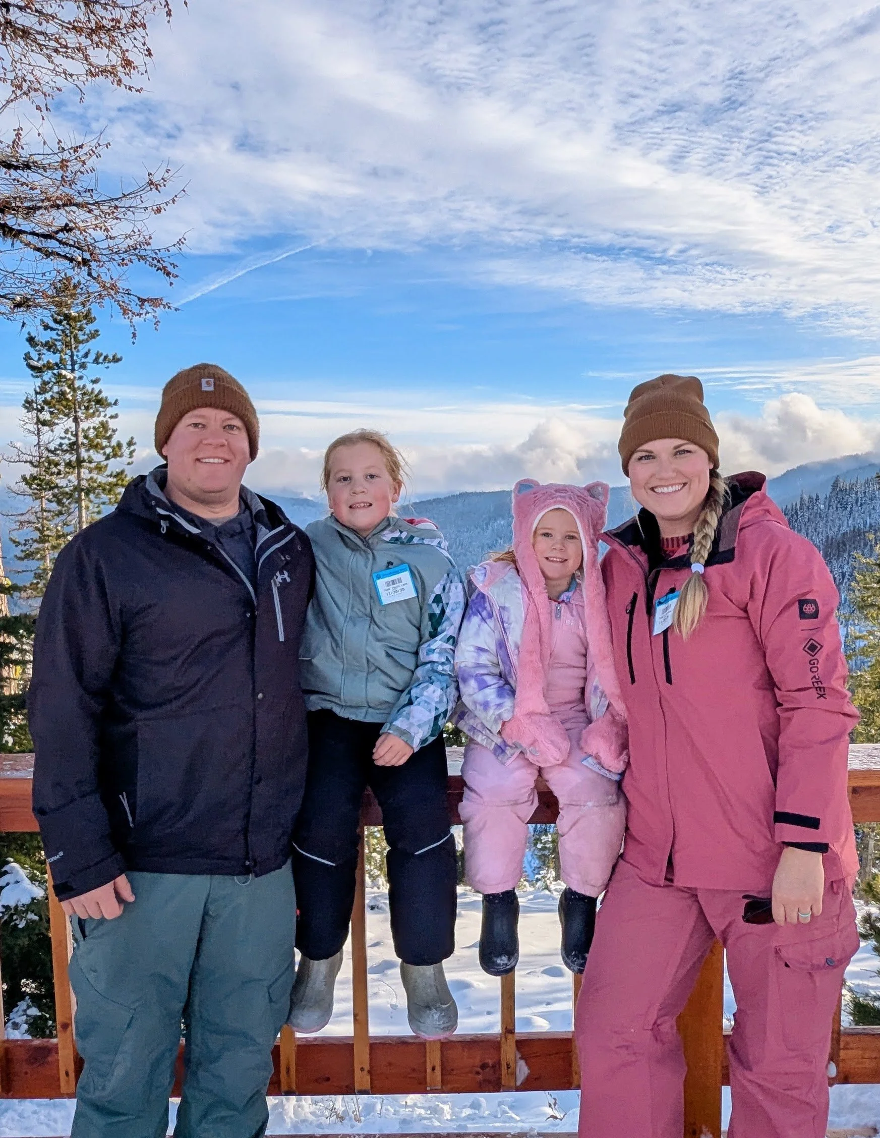 Family of four in winter clothing standing outdoors with snow on the ground and trees, smiling for a photo against a scenic mountain and cloudy sky background.