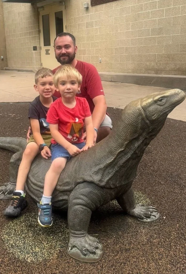 A man and two young boys sitting on a large iguana statue outdoors.