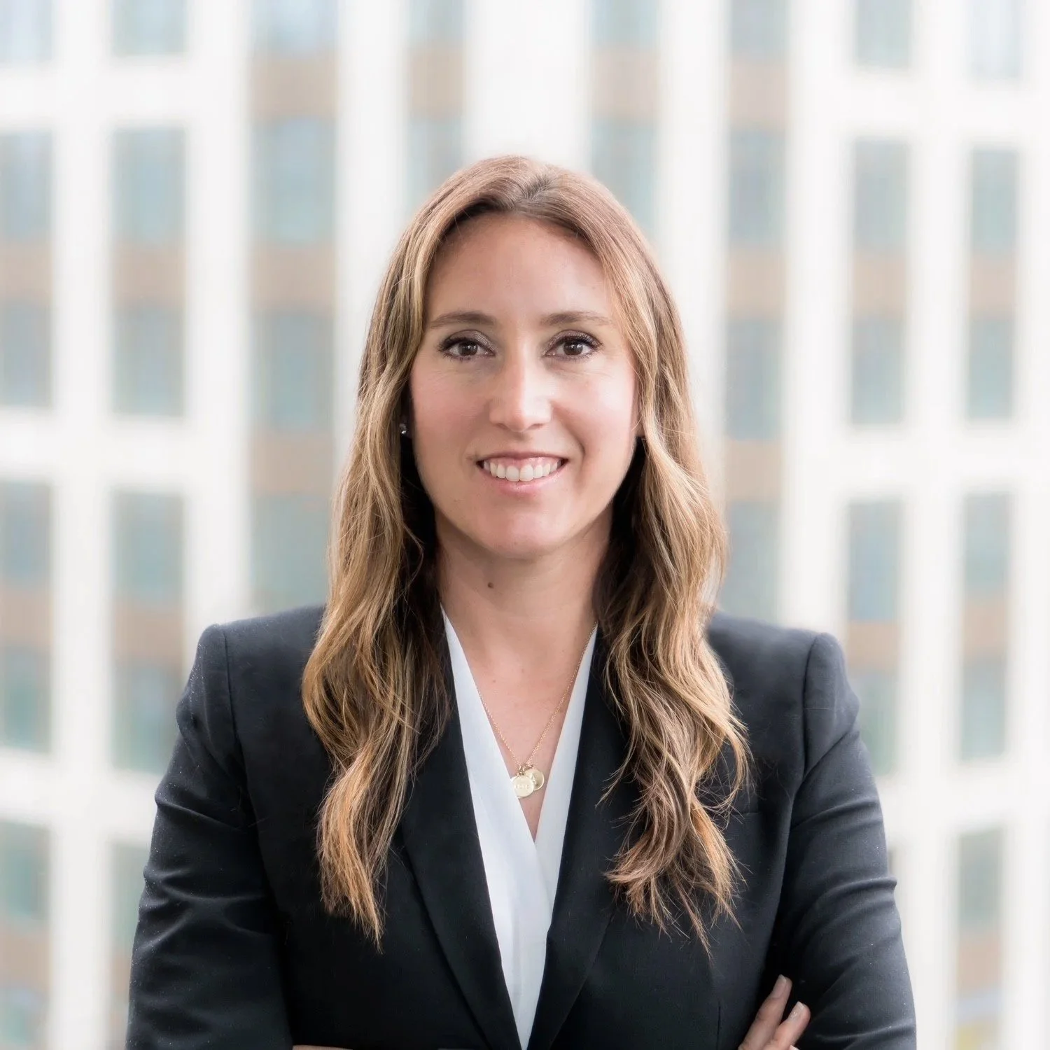 A woman with long hair wearing a black blazer and white shirt, smiling, with a background of blurred tall buildings.