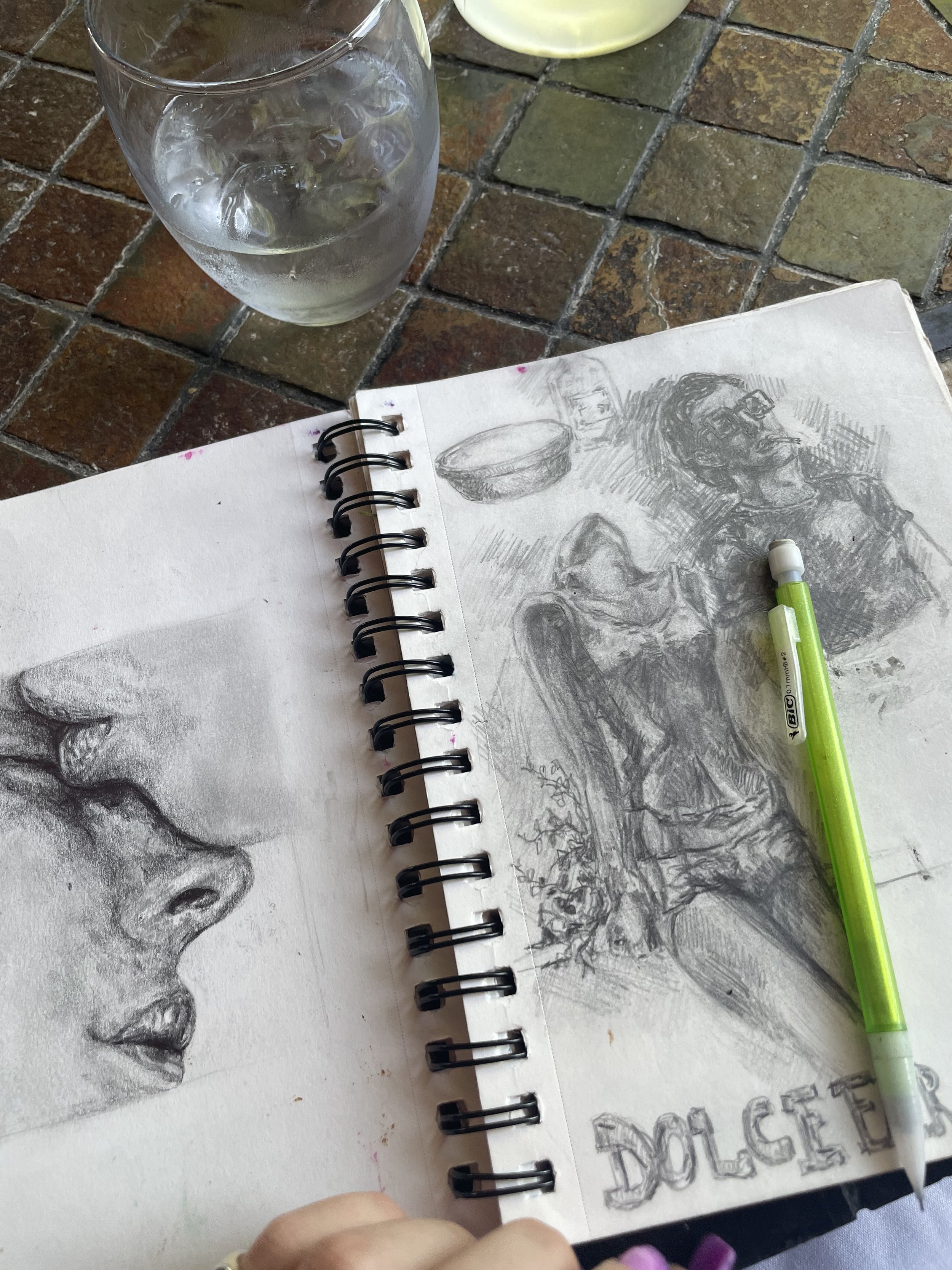 A sketchbook with detailed pencil drawings of a face and various objects, a green pencil resting on the open pages, a glass of ice water, and a partial view of a hand with painted nails on a tiled surface.