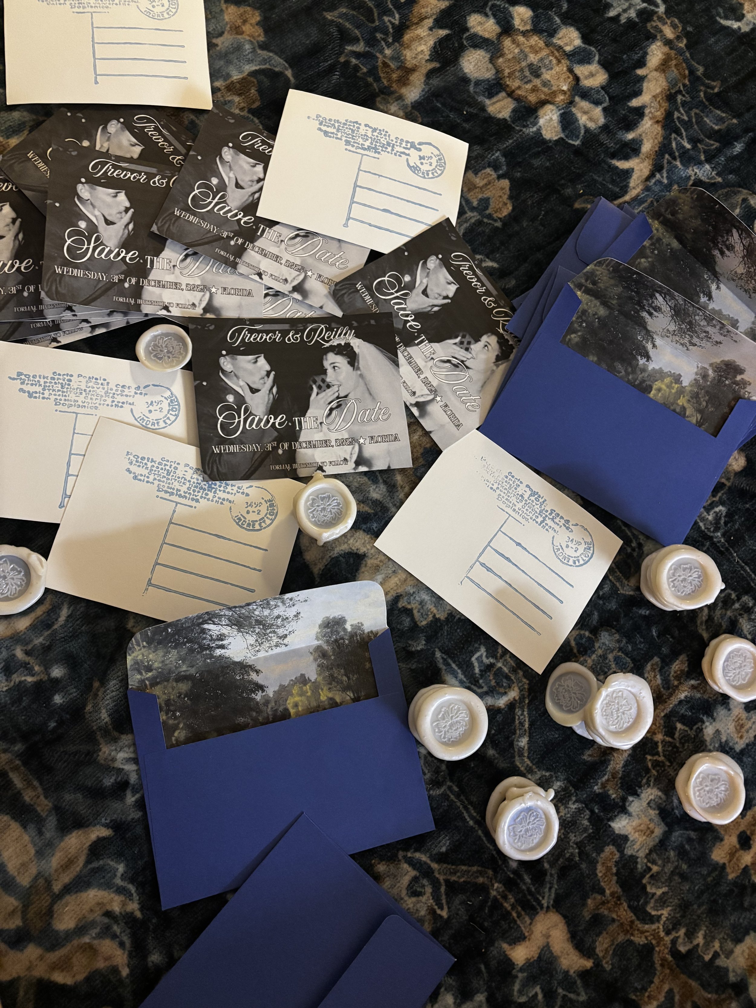 Wedding save the date cards with black and white images of a bride and groom, blue envelopes, and white wax seals with floral patterns, scattered on a dark patterned fabric.
