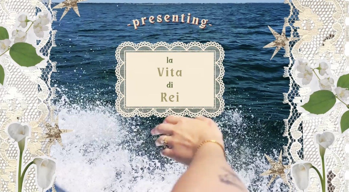 A person’s hand and arm extending over the water with ocean waves in the background, surrounded by decorative white lace and floral elements, with the text "presenting la Vita di Rei" in the center.