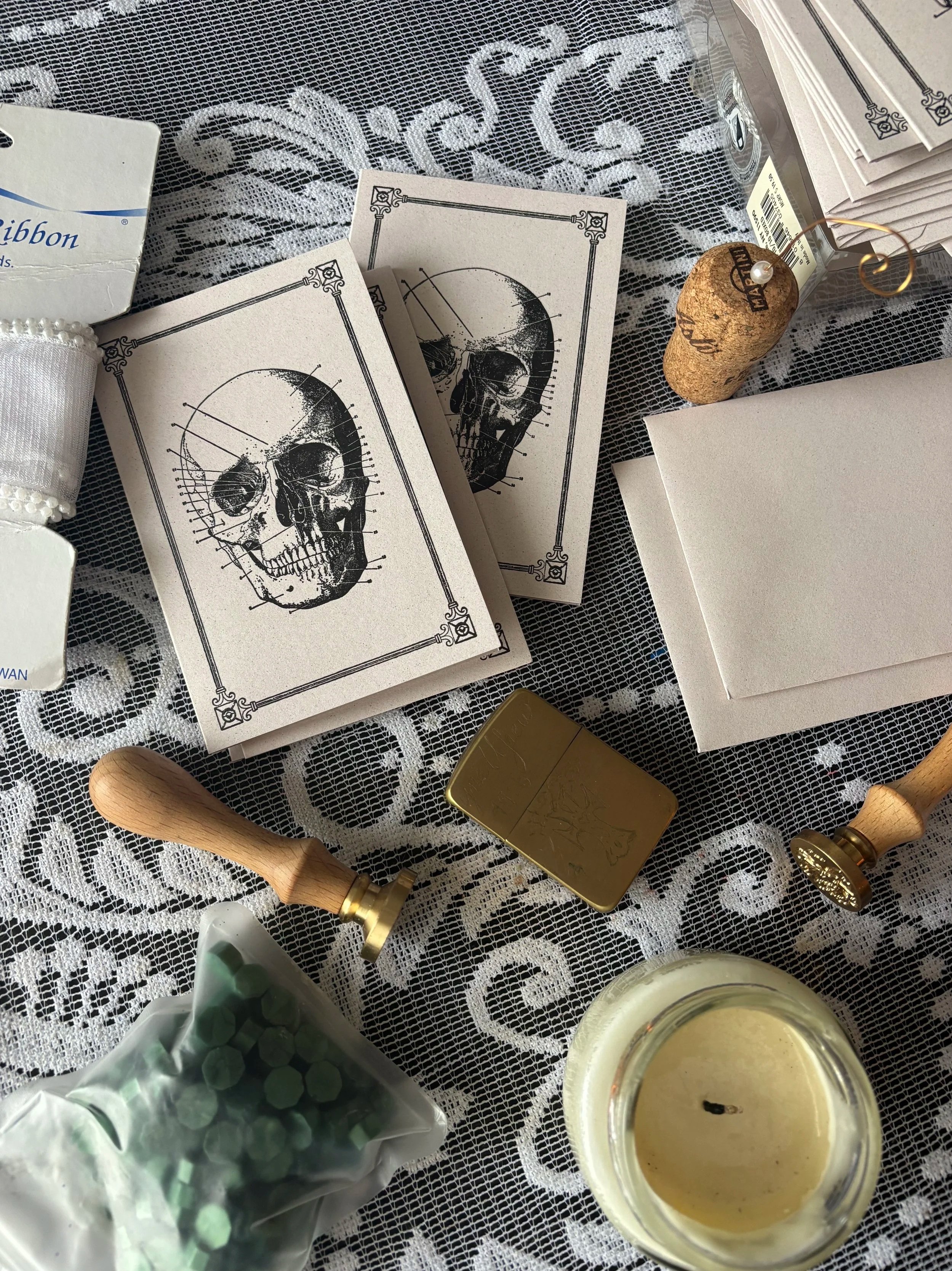 A lace tablecloth with decorative items including stationery with Italian skull illustrations, a cork topped with a pearl pin, a gold-colored tin box, a lit candle, a wooden stamp, and a bag of green wax seal beads.
