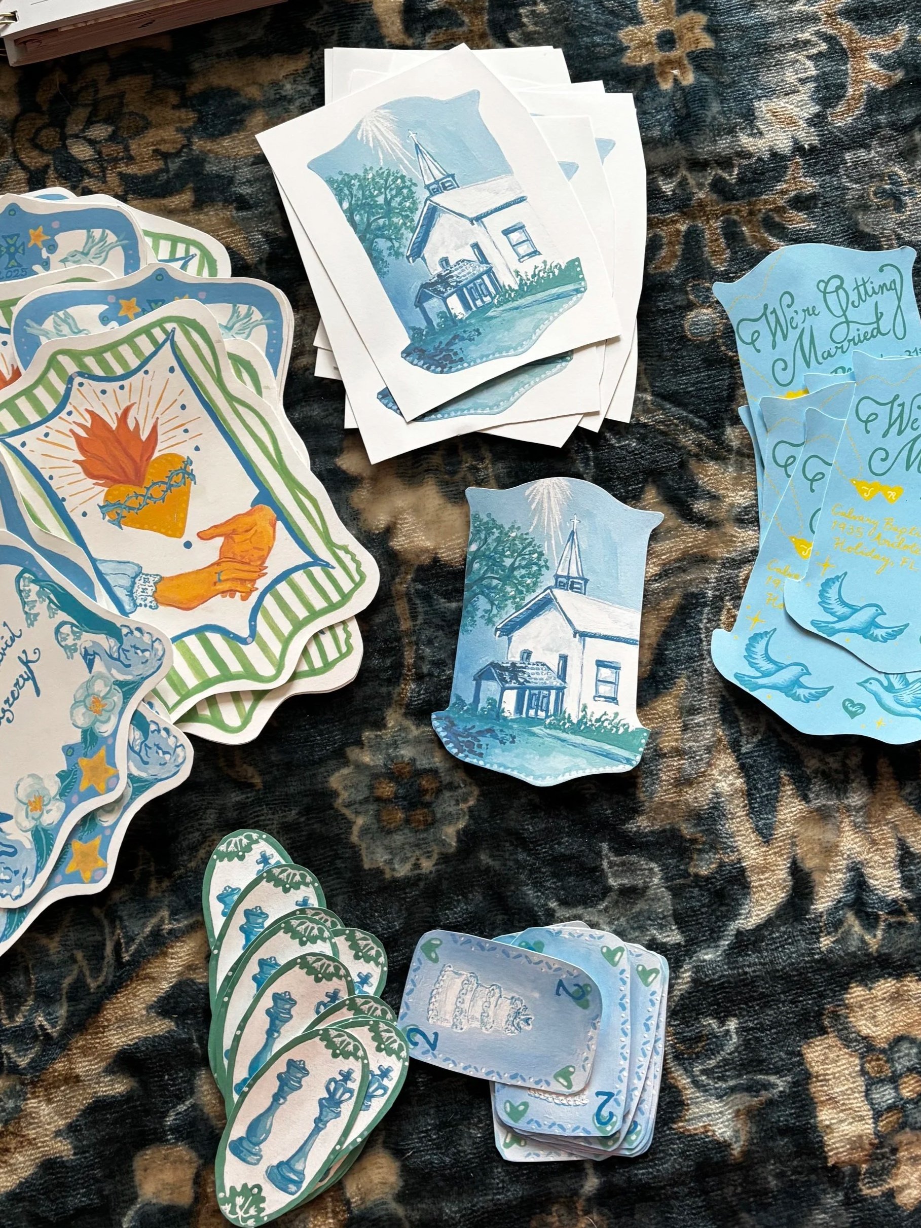 Homemade wedding stationery with illustrations of a church, a statue, and the phrase 'We're Getting Married' with a date, spread out on a patterned fabric surface.