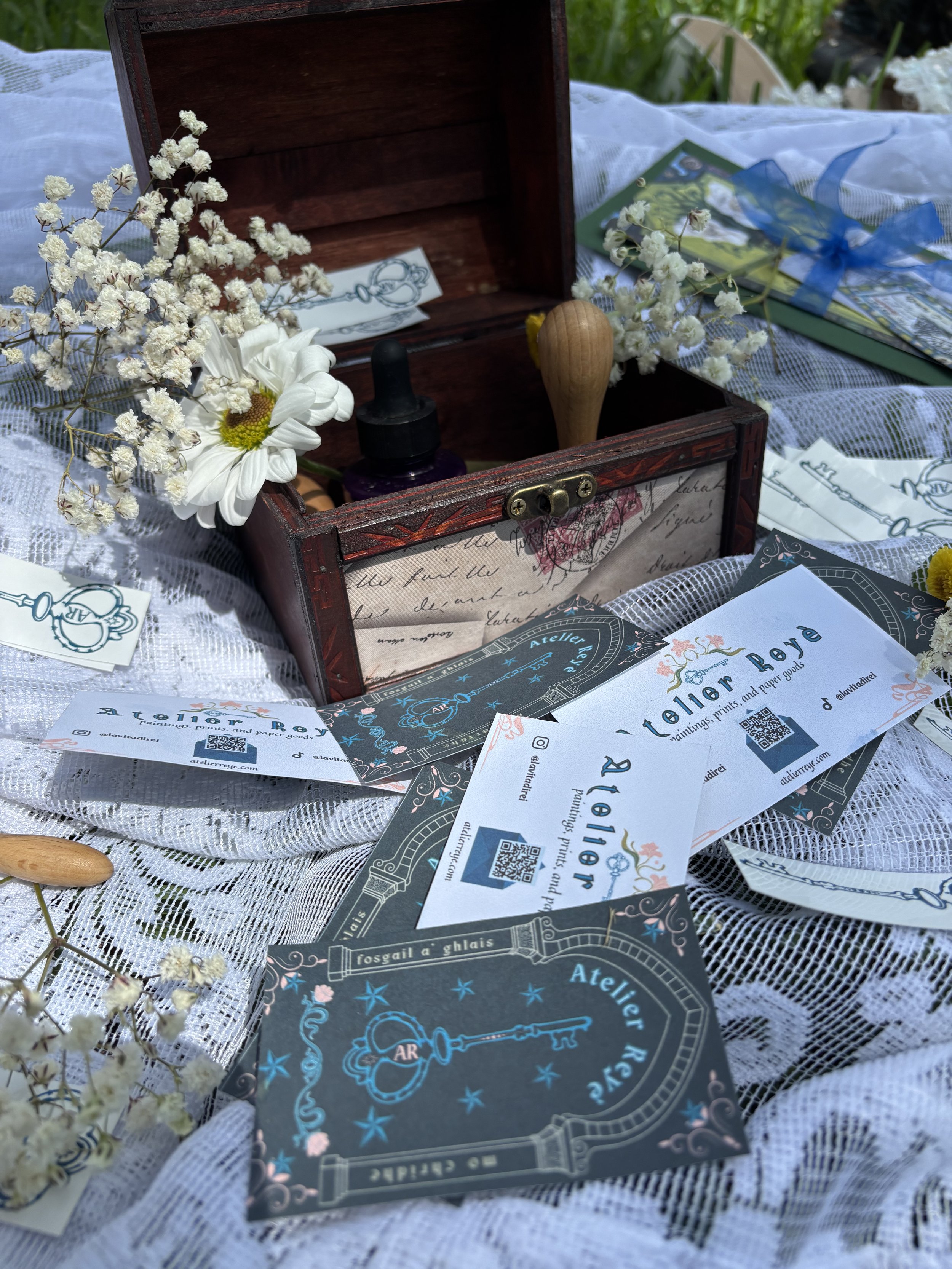 A decorated wooden box containing white flowers, small bottles, and a wax seal spoon, surrounded by business cards for Atelier Reyé on a lace tablecloth outdoors.