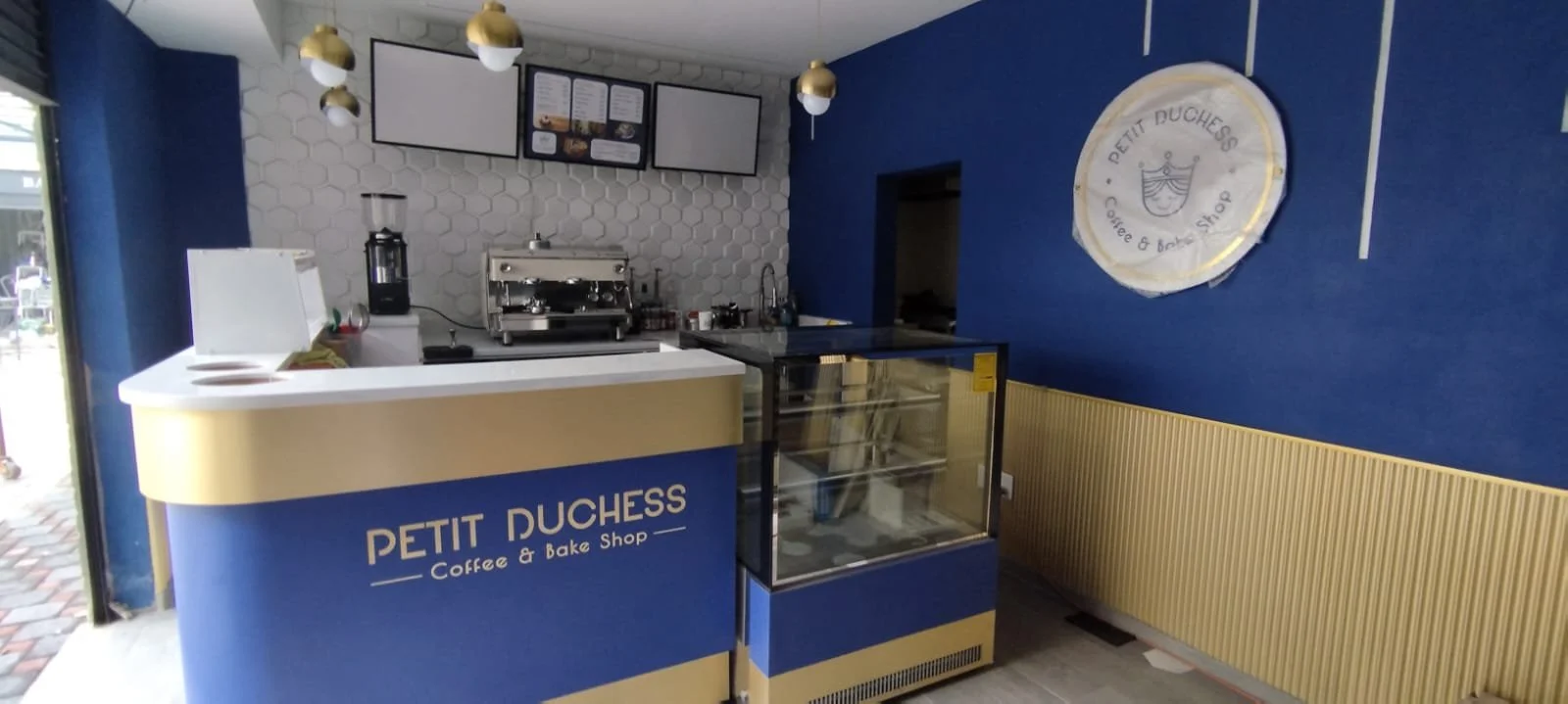 Interior of Petit Duchess Coffee & Bake Shop, featuring a blue and yellow counter, a white hexagonal tiled wall behind the counter, a coffee machine, snack display case, and a circular sign on the blue wall with the shop's name and logo.