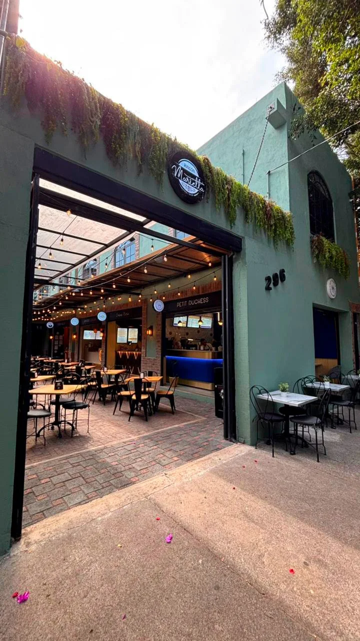 Exterior view of a restaurant with a green facade, outdoor seating with black chairs and white tables, string lights, and a sign that reads "Marcell" on the building.