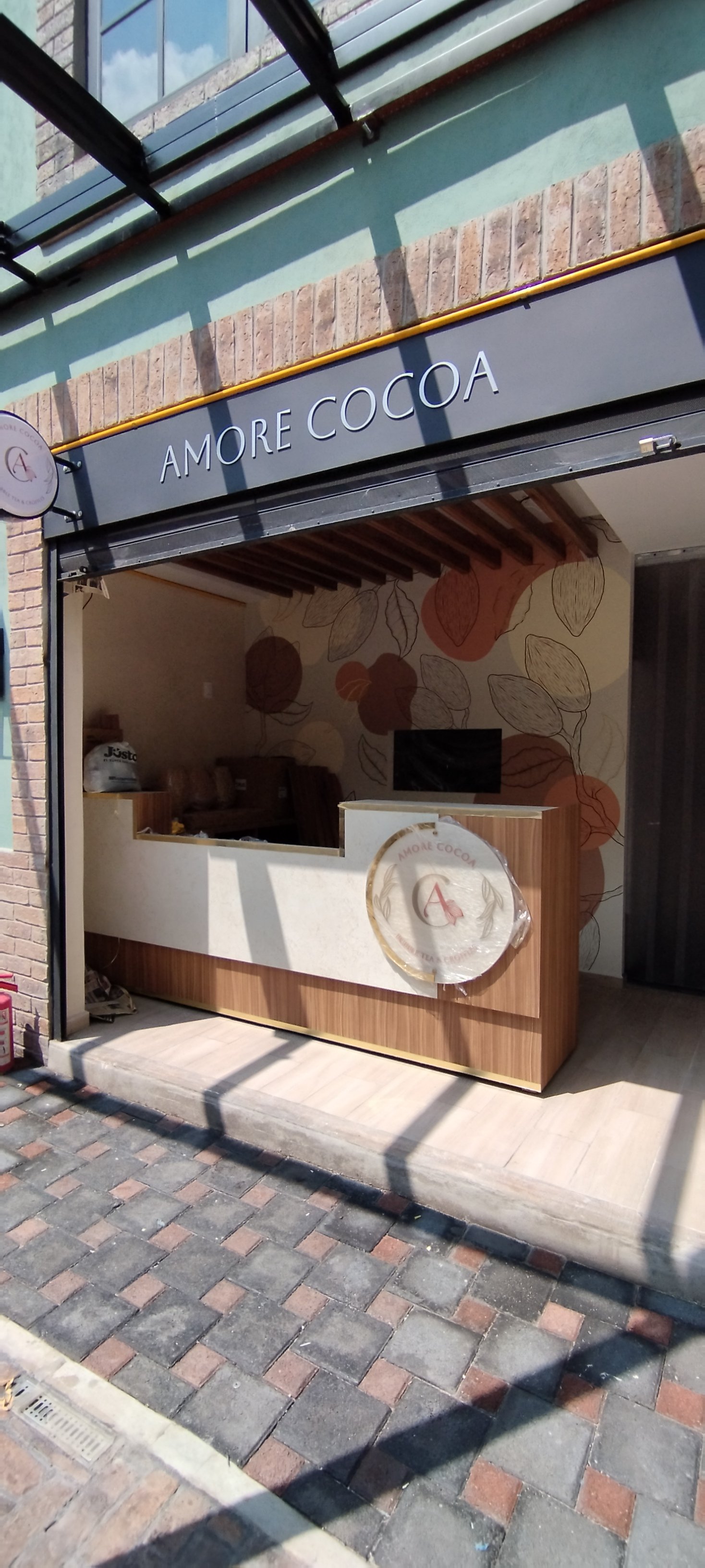 Front view of a storefront with a sign that reads 'Amore Cocoa'. The interior has a wall with an abstract leaf pattern, a counter, and a small television or monitor. The sidewalk has a brick pattern, and a fire extinguisher is visible to the left outside.